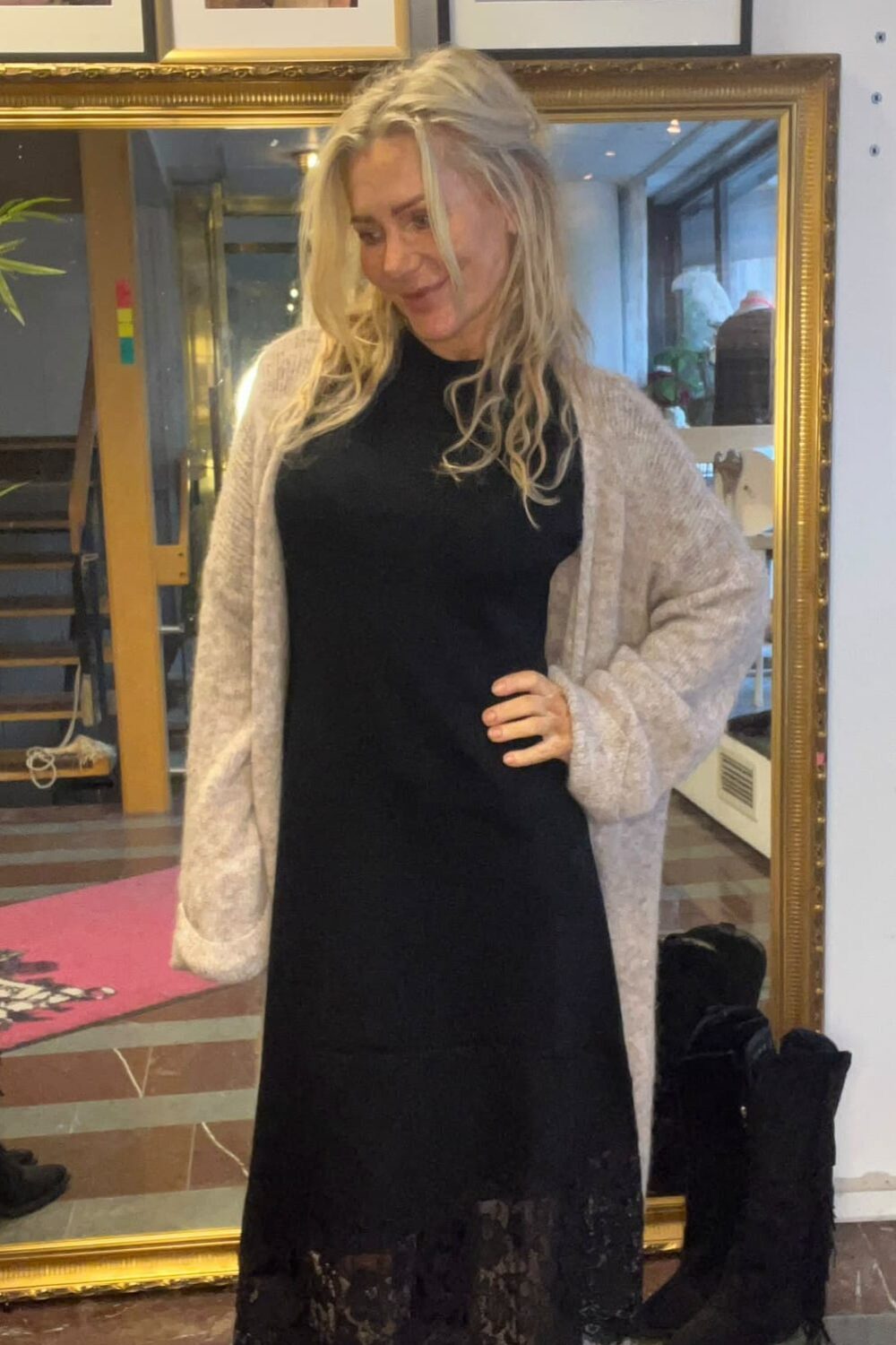 Blond kvinna poserar med handen på höften och ler lätt, iklädd svart klänning med spetskant, ljus kofta och vita stövlar. Hon står framför en stor guldramsspegel i ett rum med inramade foton på väggen.