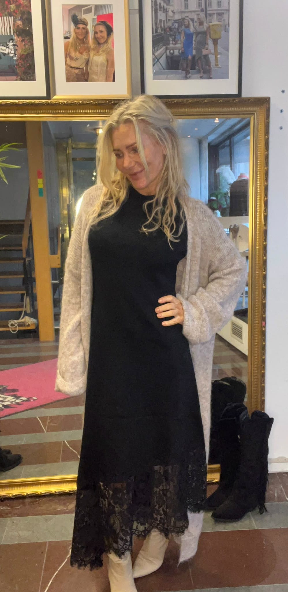 Blond kvinna poserar med handen på höften och ler lätt, iklädd svart klänning med spetskant, ljus kofta och vita stövlar. Hon står framför en stor guldramsspegel i ett rum med inramade foton på väggen.