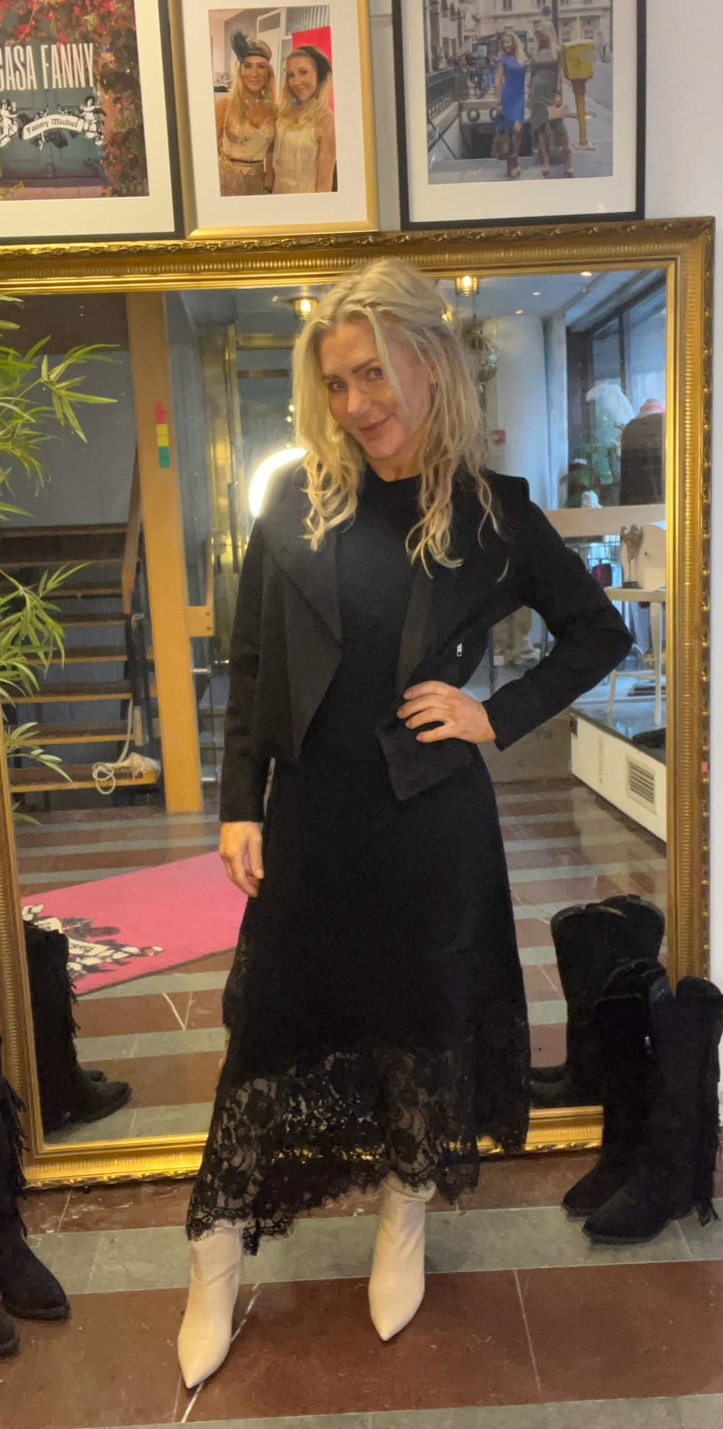 Blond kvinna poserar med handen på höften framför en stor guldramad spegel, klädd i svart klänning med spetskant och vitstövlar. I bakgrunden syns trappa, ljusslinga, växter och inramade foton på väggen.