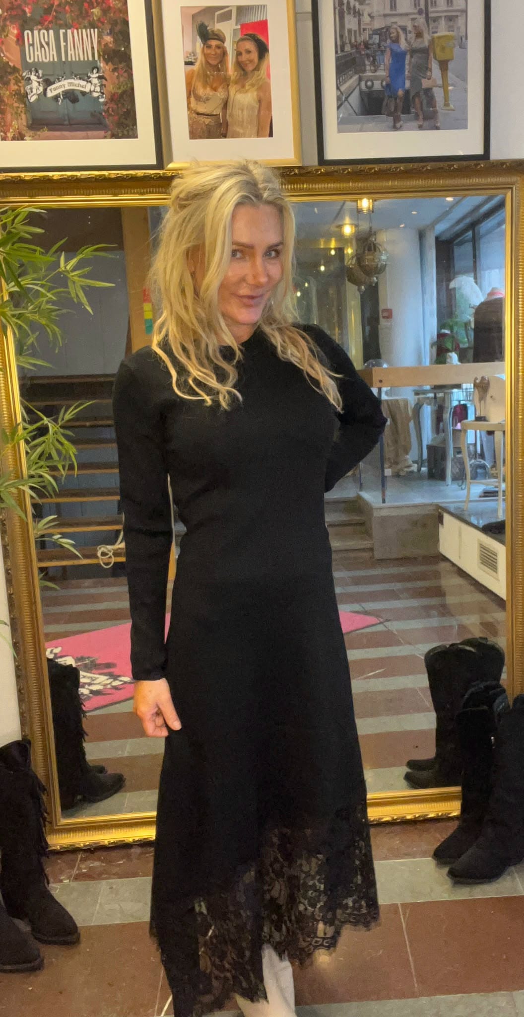 Blond kvinna poserar framför en stor guldramsspegel i en butik eller studio, klädd i lång svart klänning med spetskant. Hon ler mot kameran; i bakgrunden syns trappa, växter, lampor och inramade foton med texten “CASA FANNY”.