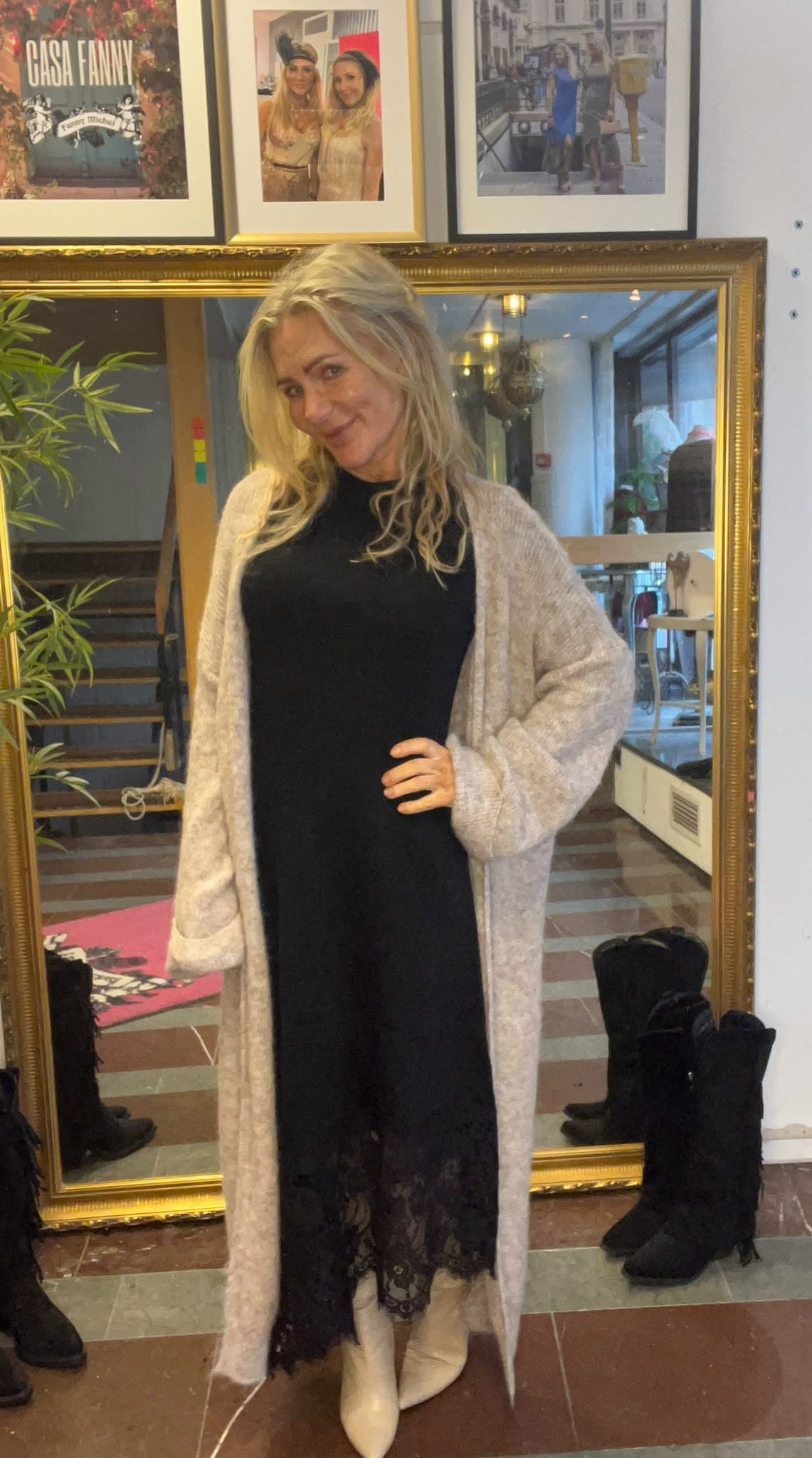 Leende blond kvinna poserar med handen på höften framför en stor guldramsspegel, iklädd svart klänning med spetskant, lång grå kofta och ljusa boots. I bakgrunden syns trappa, växter och inramade foton. Text: “CASA FANNY”.