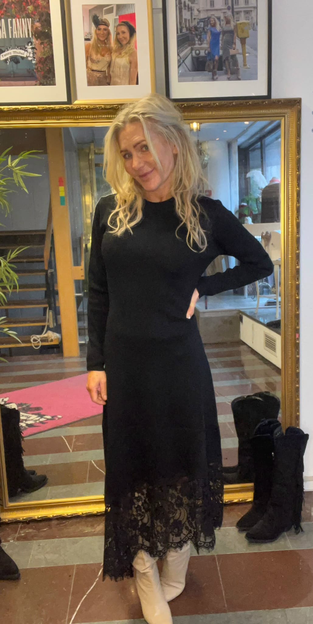 Blond kvinna i lång svart klänning med spetskant står och ler med handen på höften, framför en stor guldramsspegel. I bakgrunden syns trappa, växt och inramade foton på väggen samt ett par svarta stövlar.