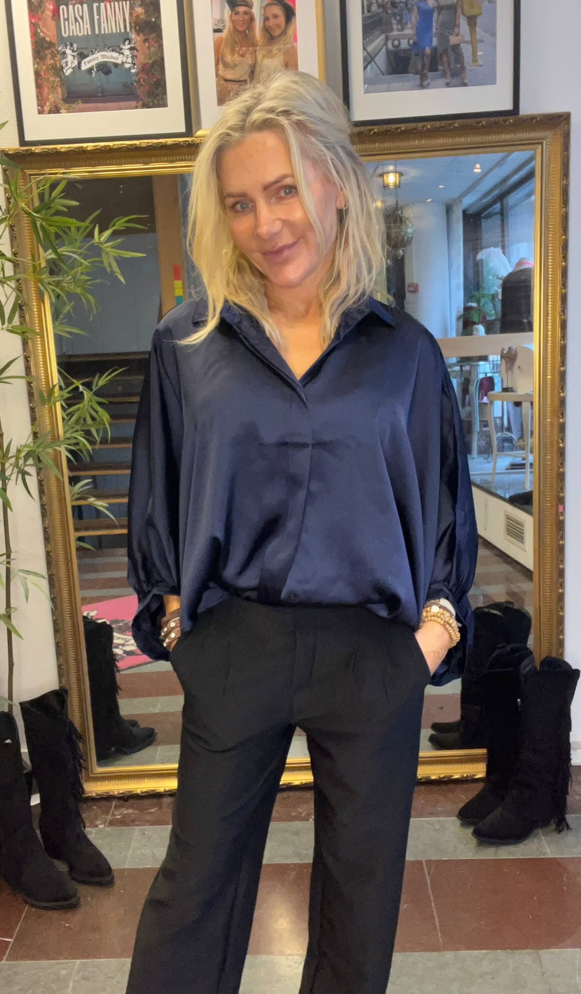 Blond kvinna står och ler mot kameran med händerna i fickorna, klädd i mörkblå blus och svarta byxor. Bakom henne syns en stor guldramsspegel, stövlar på golvet och inramade foton, inklusive texten “CASA FANNY”.