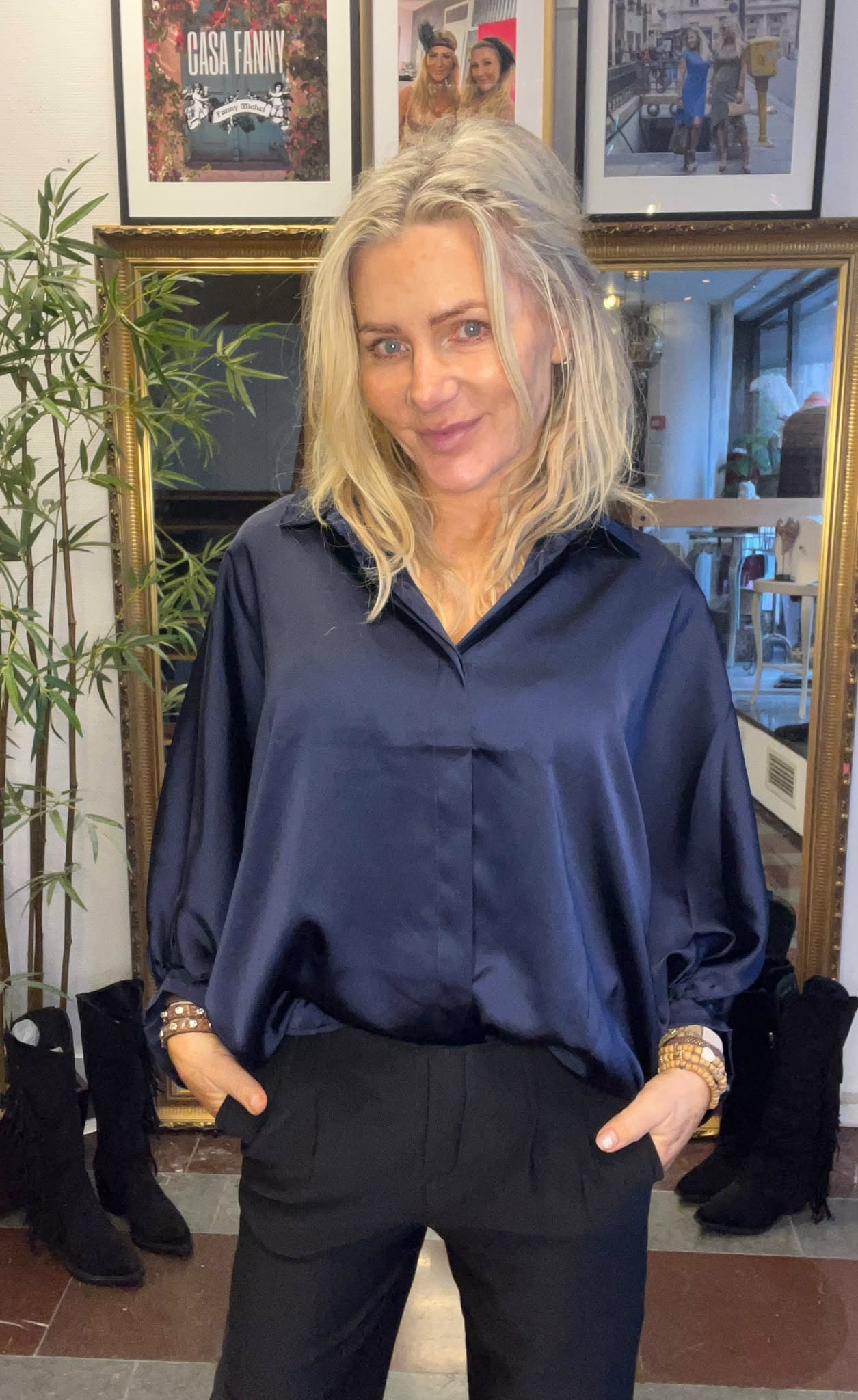 Blond kvinna står och ler med händerna i fickorna, klädd i mörkblå blus och svarta byxor. Bakom henne syns en guldramsspegel, en grön växt och inramade foton; ett med texten ”CASA FANNY”.