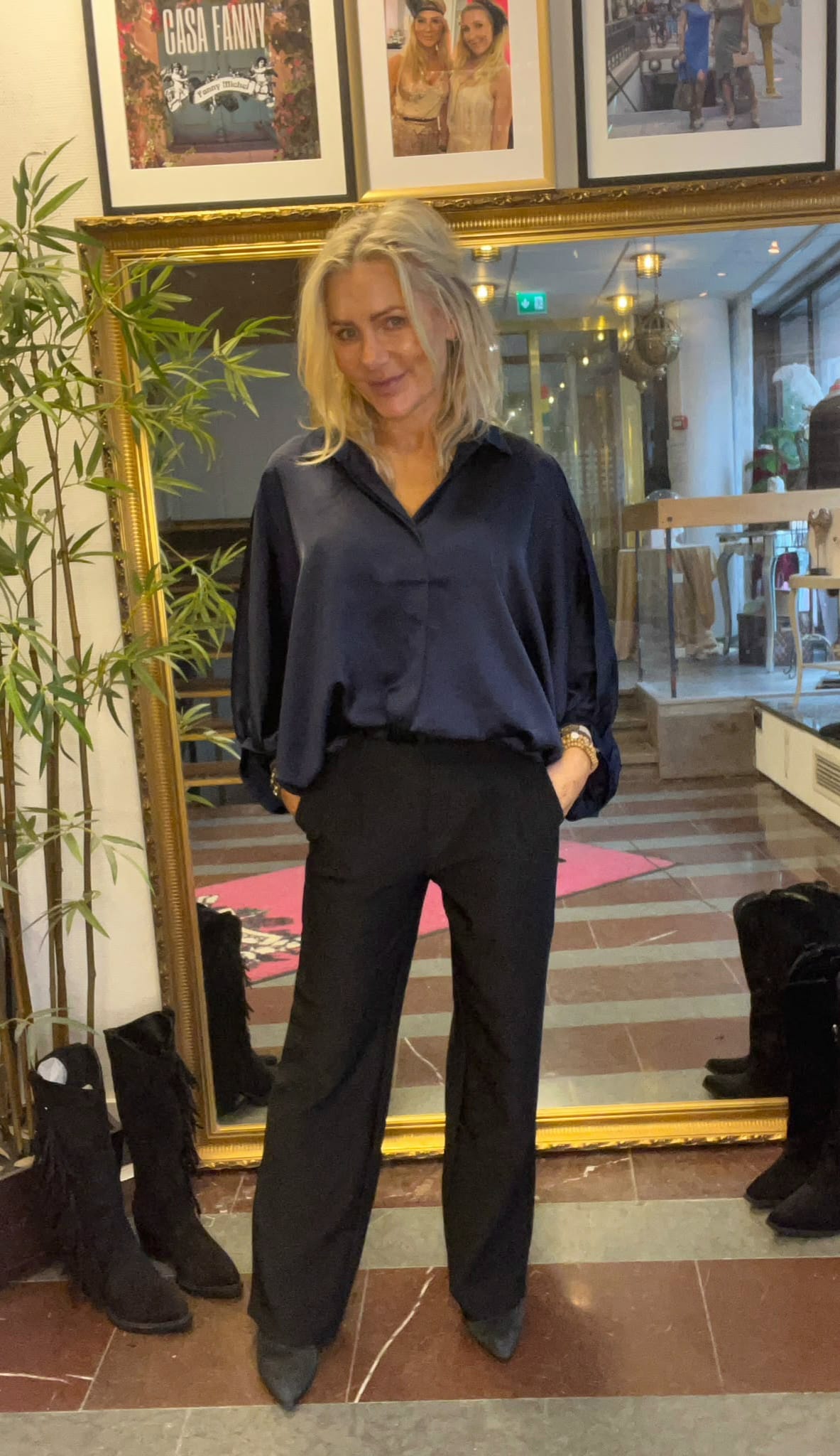 Blond kvinna står avslappnat med händerna i fickorna framför en stor guldramsspegel i en hall. Hon bär mörkblå blus, svarta byxor och svarta skor; växt, boots och inramade foton syns runtom. Text: “CAISA FANNY”.