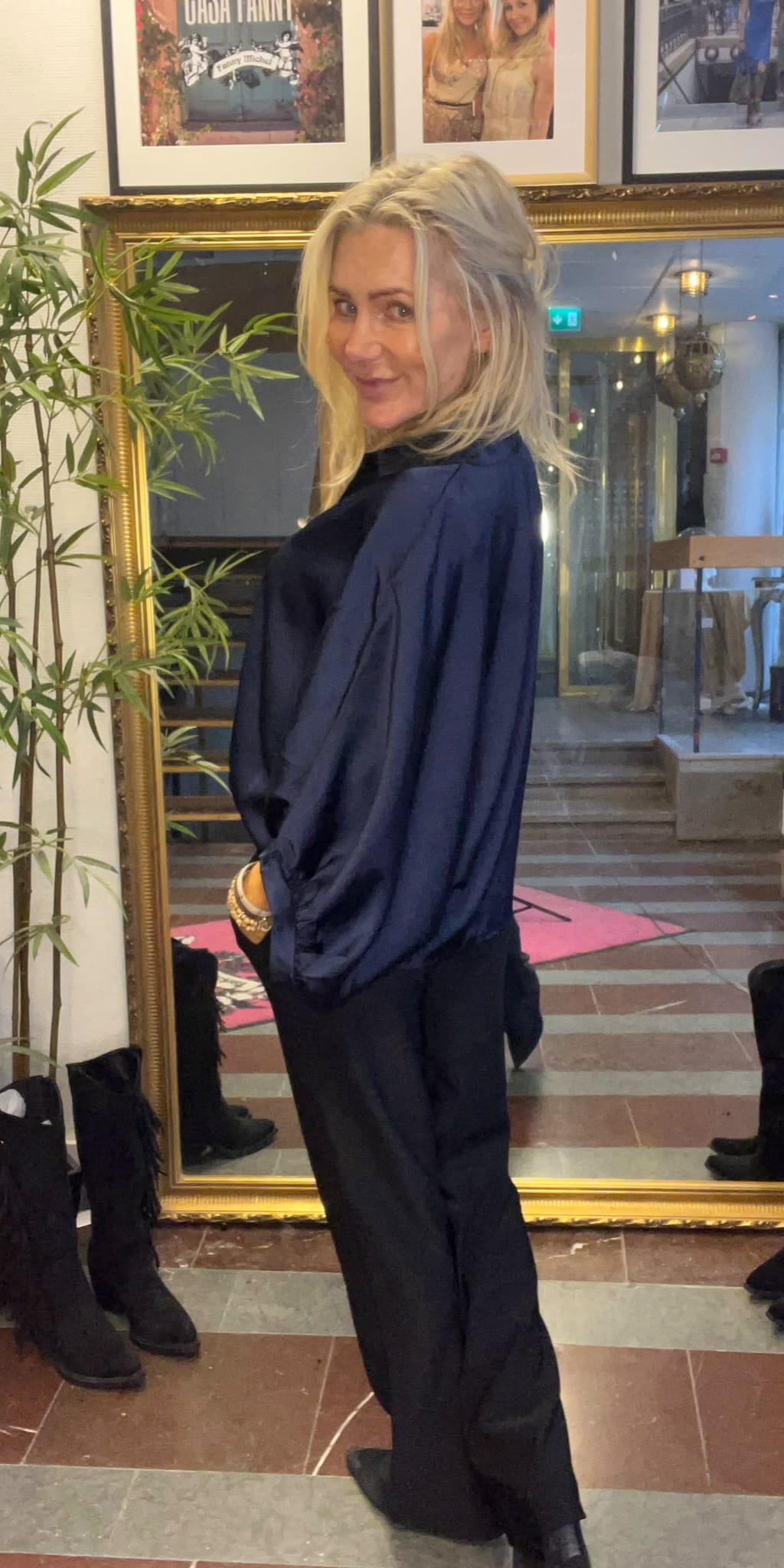 Blond kvinna i mörkblå blus och svarta byxor står i profil med händerna i fickorna och ler mot kameran. Hon står framför en stor guldramsspegel i en hall med foton på väggen.
