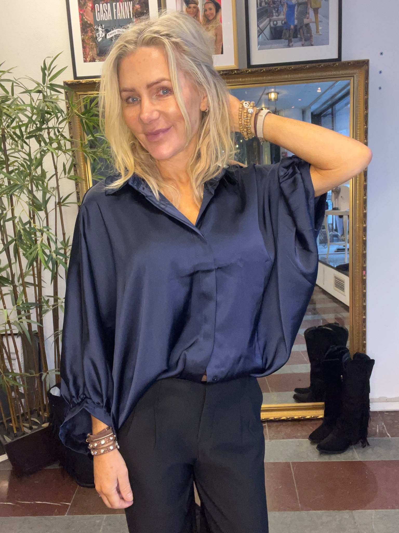 Blond kvinna i mörkblå blus och svarta byxor poserar och ler, med ena armen bakom huvudet. Hon står inomhus framför en stor guldramsspegel och väggkonst med texten “CASA FANNY”.
