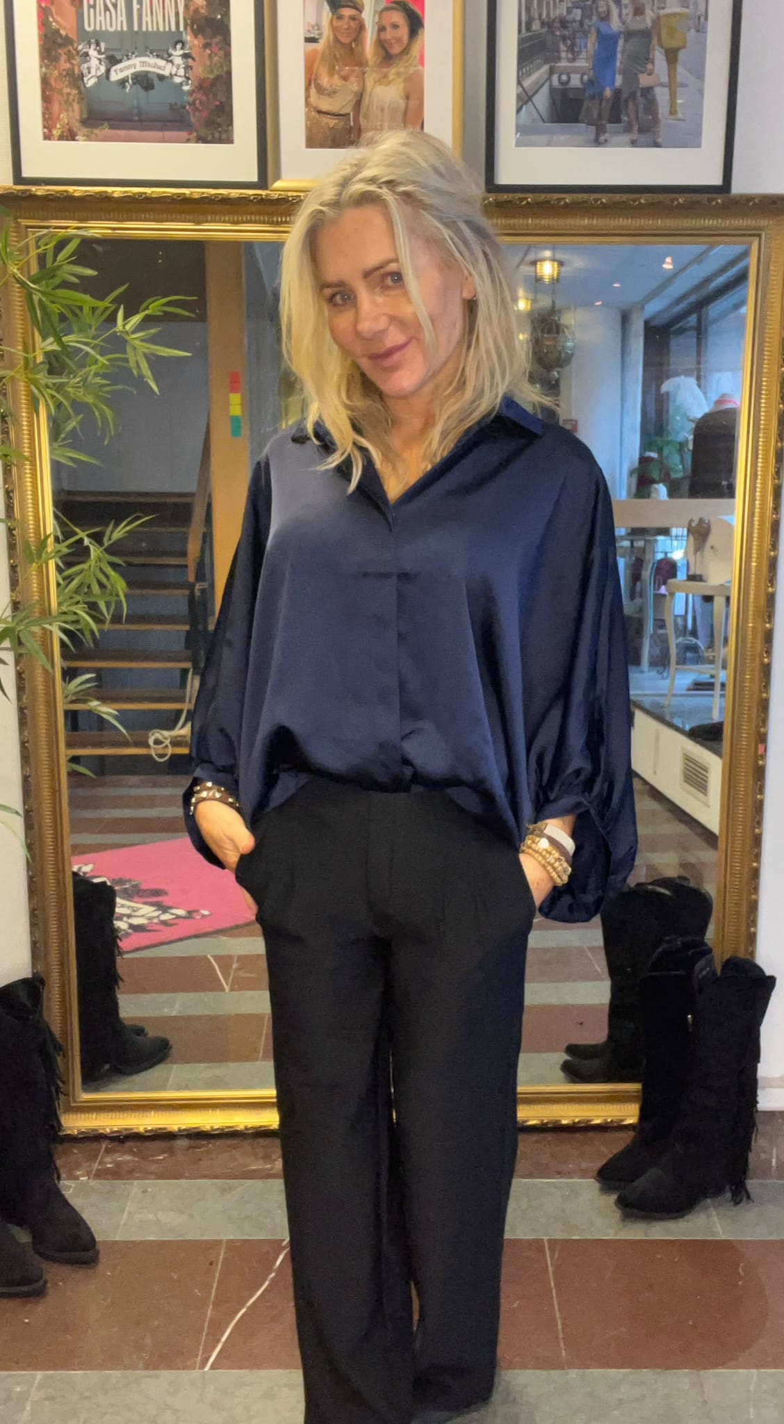 Blond kvinna står och ler med händerna i fickorna, klädd i mörkblå blus och svarta byxor, framför en stor guldramsspegel i en hall. I bakgrunden syns bambuplanta, trappa och inramade foton med texten “CHISA FANNY”.