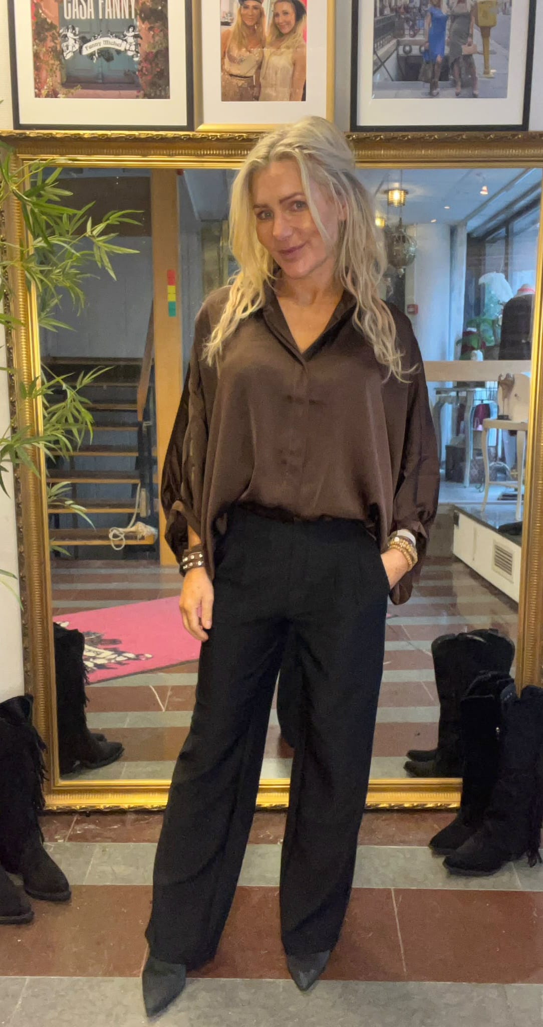 Blond kvinna står och poserar med ena handen i fickan framför en stor guldramsspegel, klädd i mörk blus och svarta byxor. I bakgrunden syns trappa, växter och inramade foton; texten “CASA FANNY”.