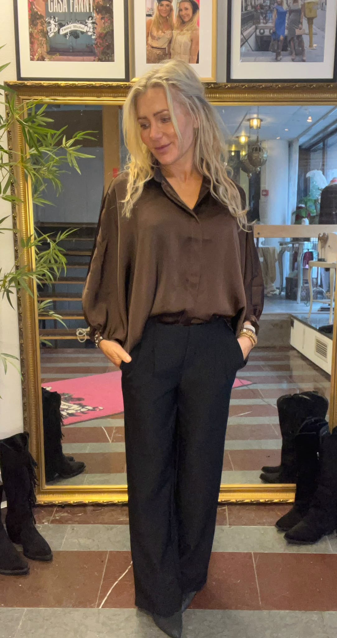 Blond kvinna står med händerna i fickorna och tittar nedåt, klädd i brun blus och svarta vida byxor. Hon poserar framför en stor guldramsspegel i en butik med inramade foton och texten “CASA FANNY”.