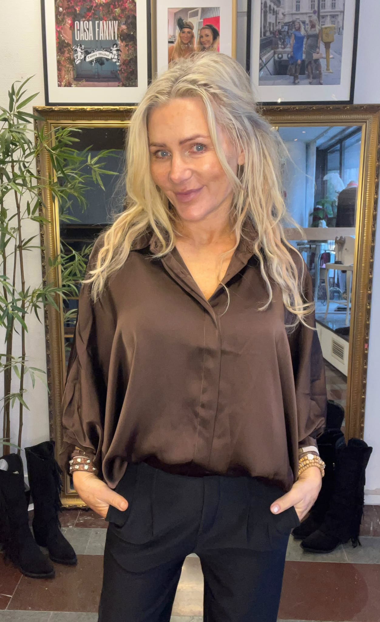 Blond kvinna står och ler med händerna i fickorna, klädd i brun blus och svarta byxor. Bakom henne syns en stor guldramsspegel, en grön växt och inramade foton; ett med texten “CASA FANNY”.