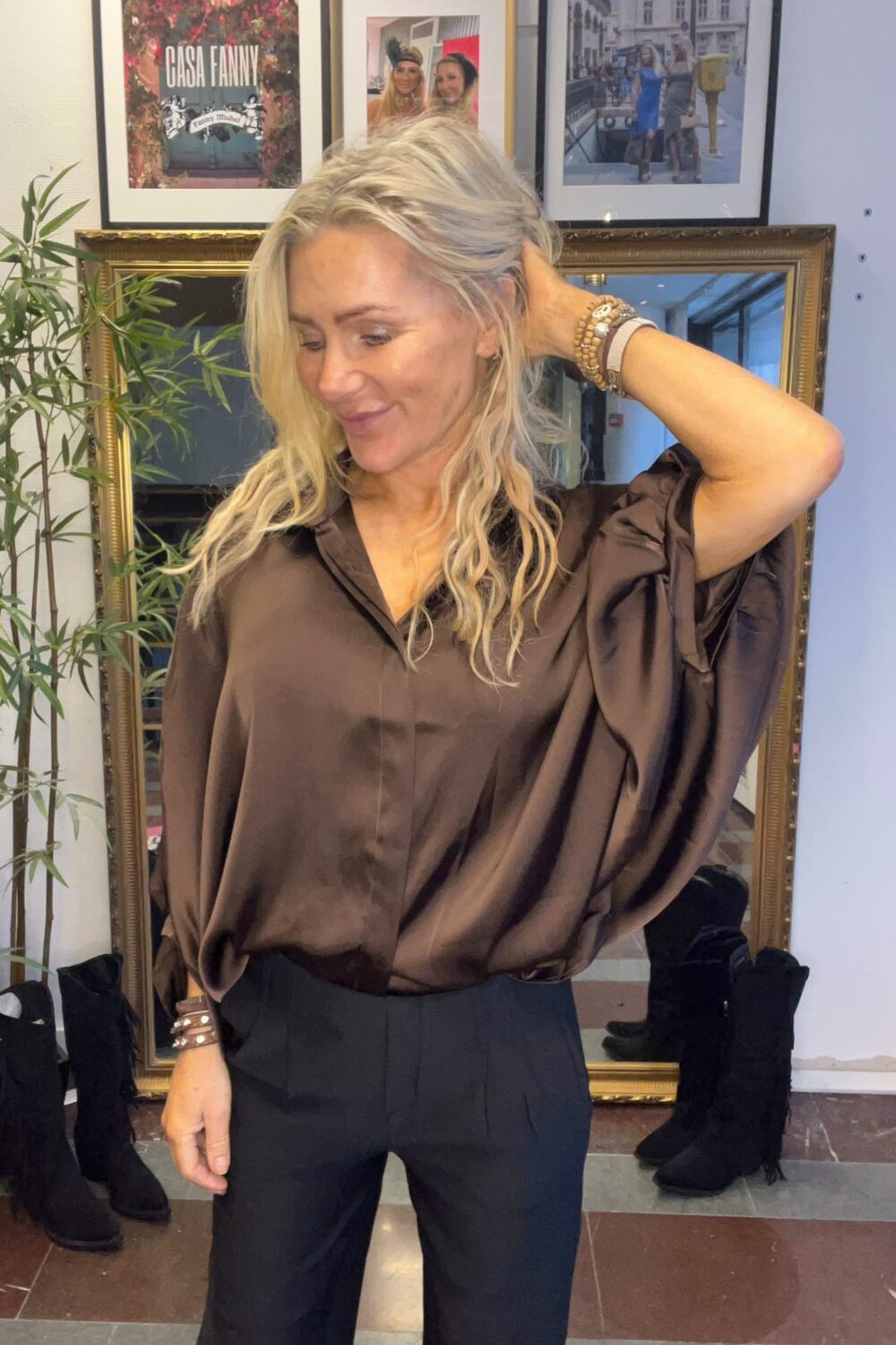 Blond kvinna poserar och drar handen genom håret, klädd i brun satinblus och svarta byxor. Hon står framför en guldramsspegel med en växt bredvid; på väggen syns inramade foton och texten ”CASA FANNY”.