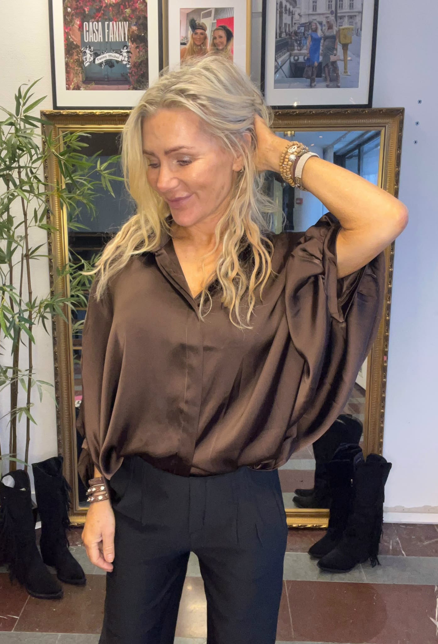 Blond kvinna poserar och drar handen genom håret, klädd i brun satinblus och svarta byxor. Hon står framför en guldramsspegel med en växt bredvid; på väggen syns inramade foton och texten ”CASA FANNY”.