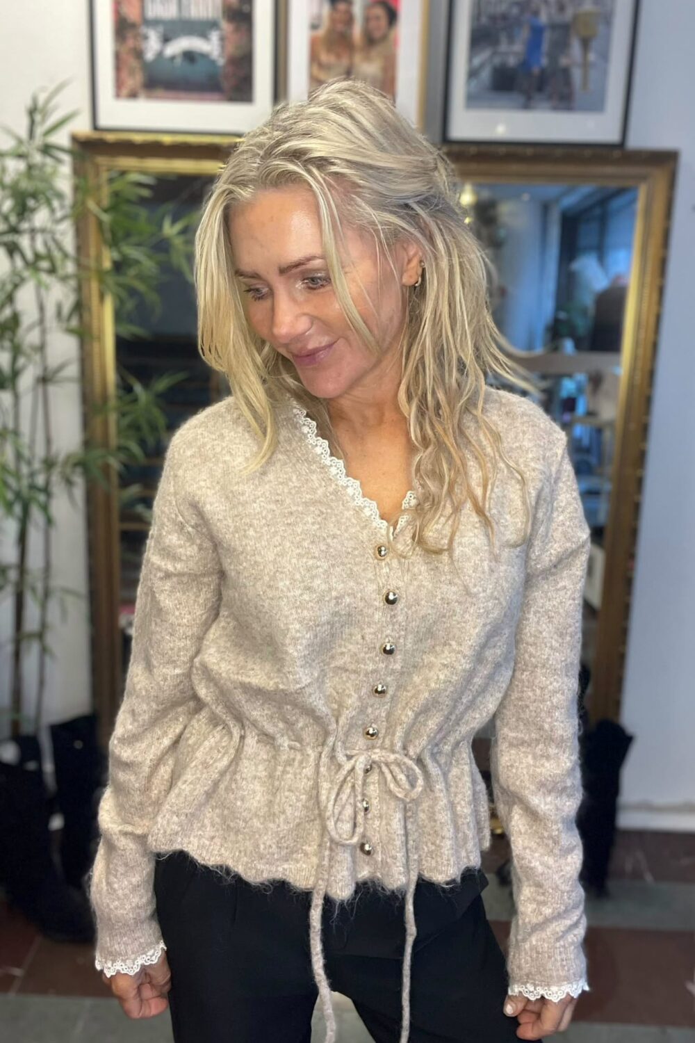Blond kvinna står inomhus och tittar ned med ett litet leende, klädd i ljusbeige stickad kofta med spetskant, knappar och knytband. I bakgrunden syns stor guldramsspegel, växt och inramade foton på väggen.