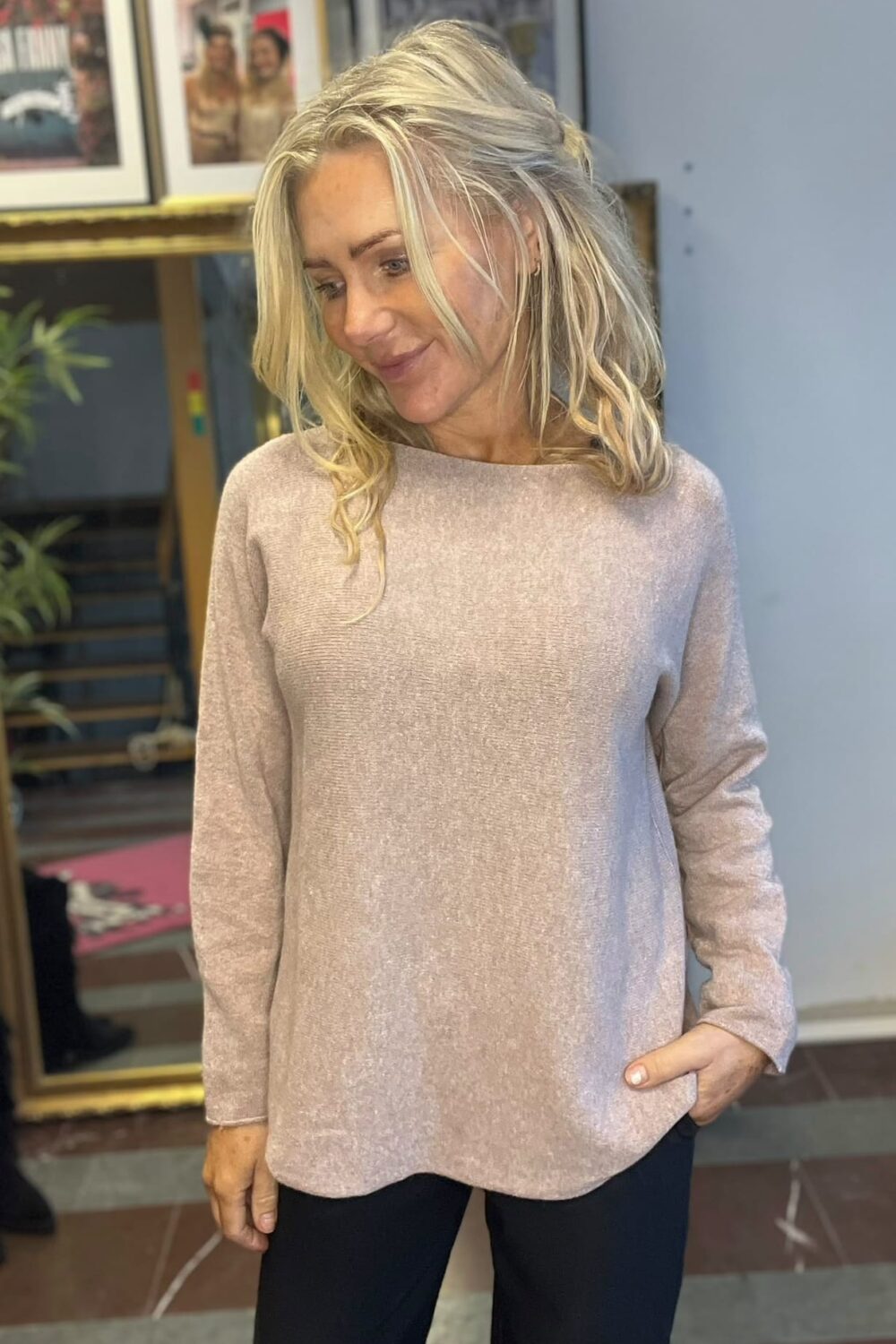 Blond kvinna står inomhus och tittar nedåt med ett svagt leende, ena handen i fickan. Hon bär en ljusrosa stickad tröja och mörka byxor; bakom syns inramade foton och en spegel.