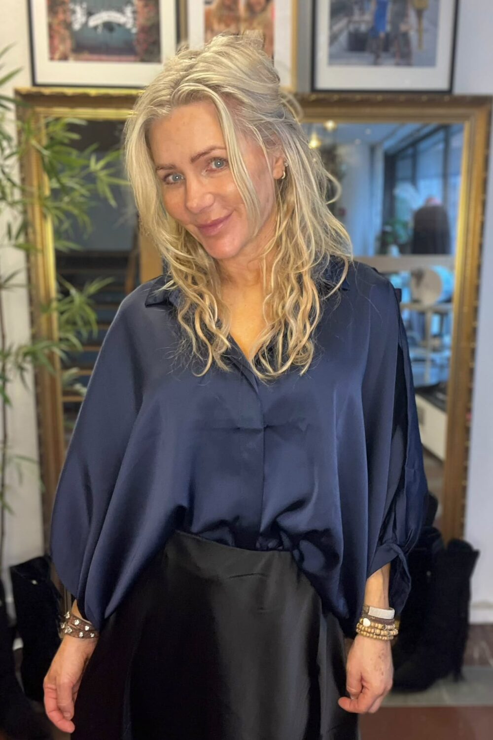 Blond kvinna ler mot kameran och står lätt vinklad, klädd i mörkblå blus och svart kjol, med armband på handlederna. Bakom syns en stor spegel, en växt och inramade foton på väggen.