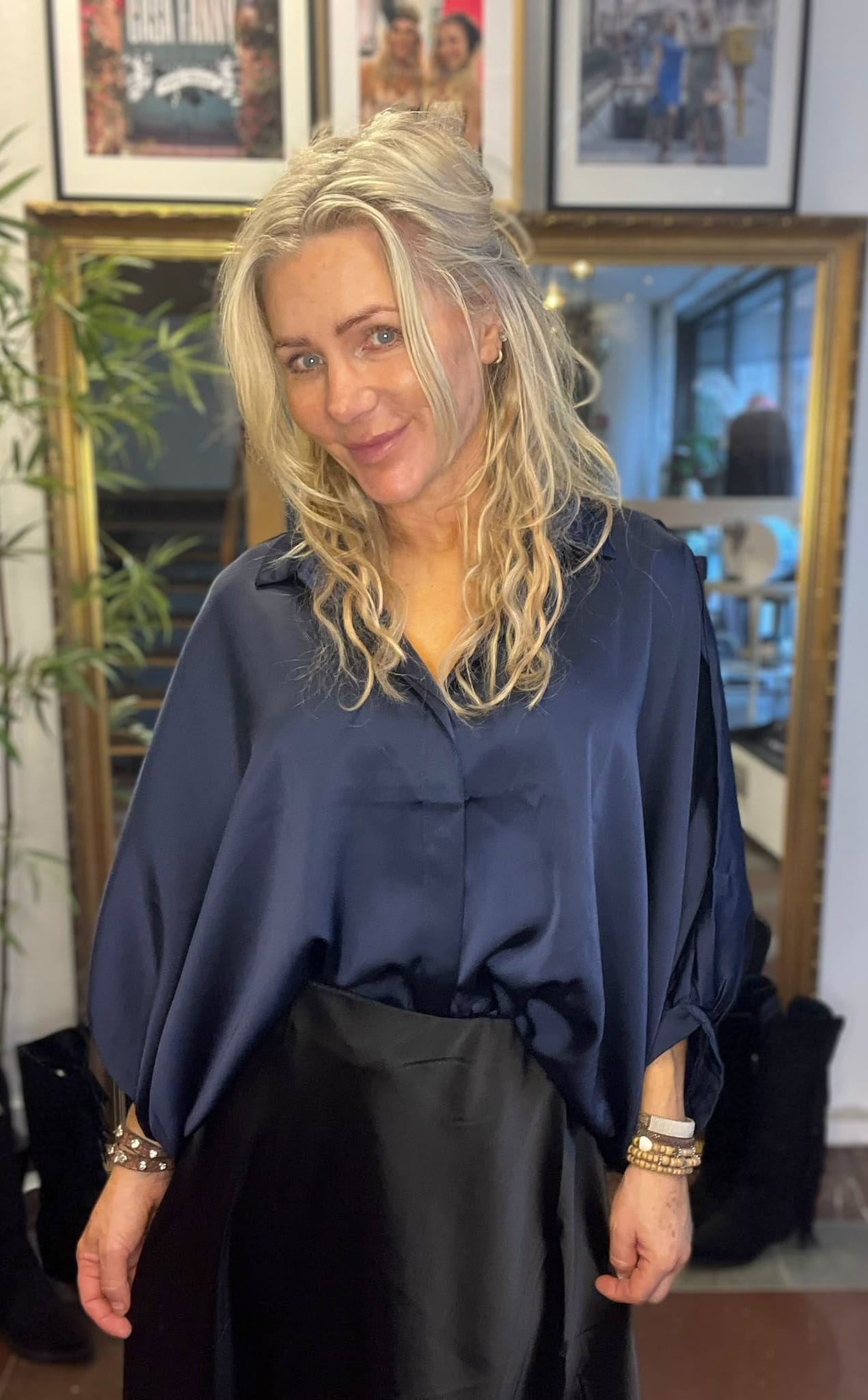 Blond kvinna ler mot kameran och står lätt vinklad, klädd i mörkblå blus och svart kjol, med armband på handlederna. Bakom syns en stor spegel, en växt och inramade foton på väggen.