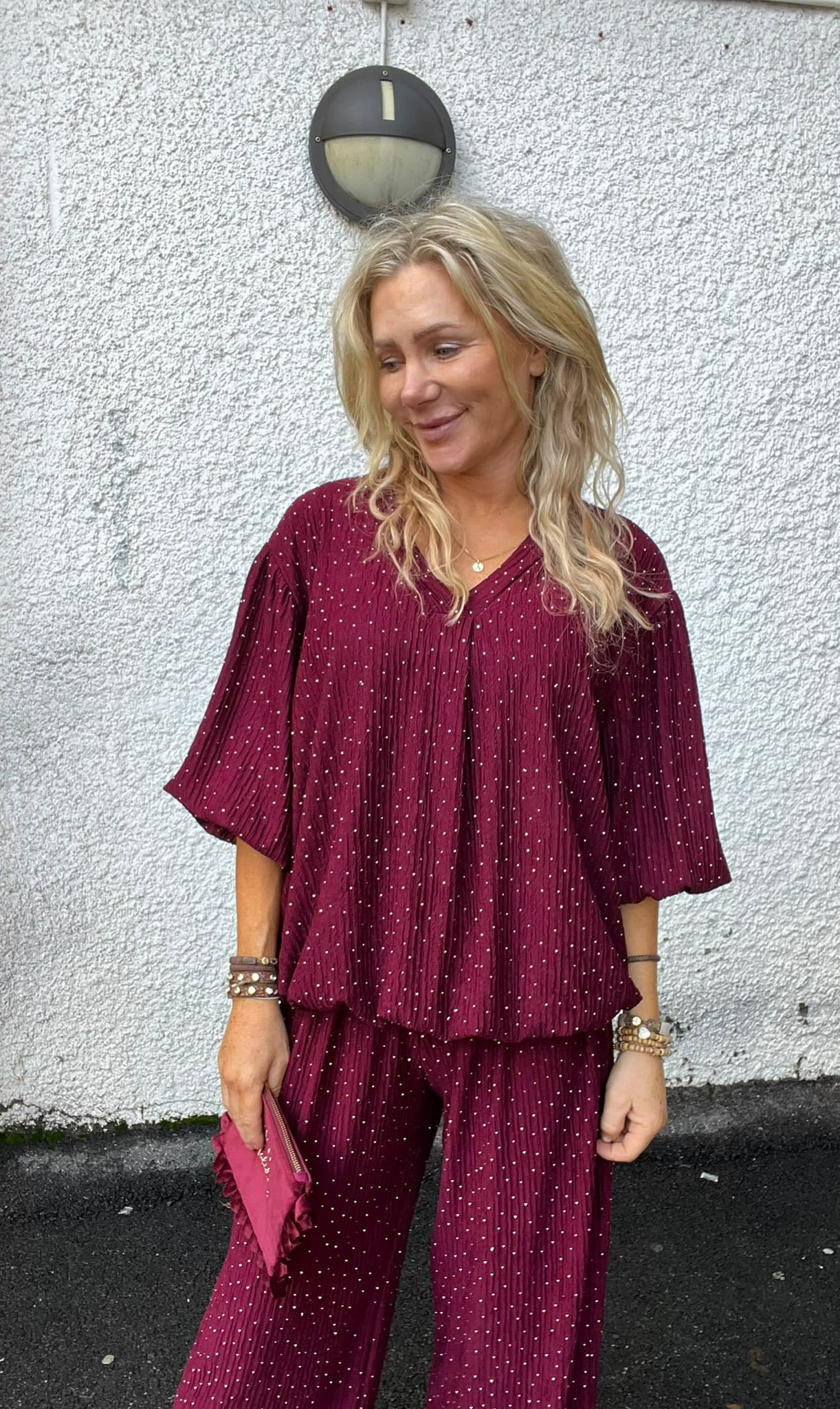 Blond kvinna står avslappnat och ler medan hon tittar åt sidan, klädd i vinröd prickig matchande topp och byxor. Hon håller en vinröd clutch och bär armband, framför en vit putsad vägg med rund vägglampa.