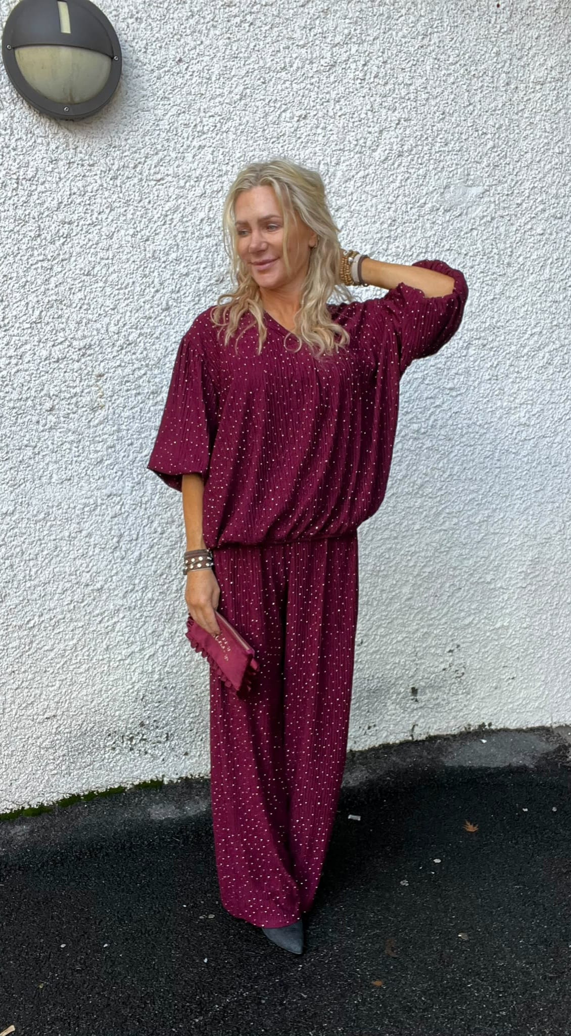 Blond kvinna står avslappnat mot en vit, grovputsad vägg och ler, med ena handen i håret. Hon bär vinröd, prickig tvådelad outfit, håller en liten clutch och har armband; vägglampa syns ovanför.