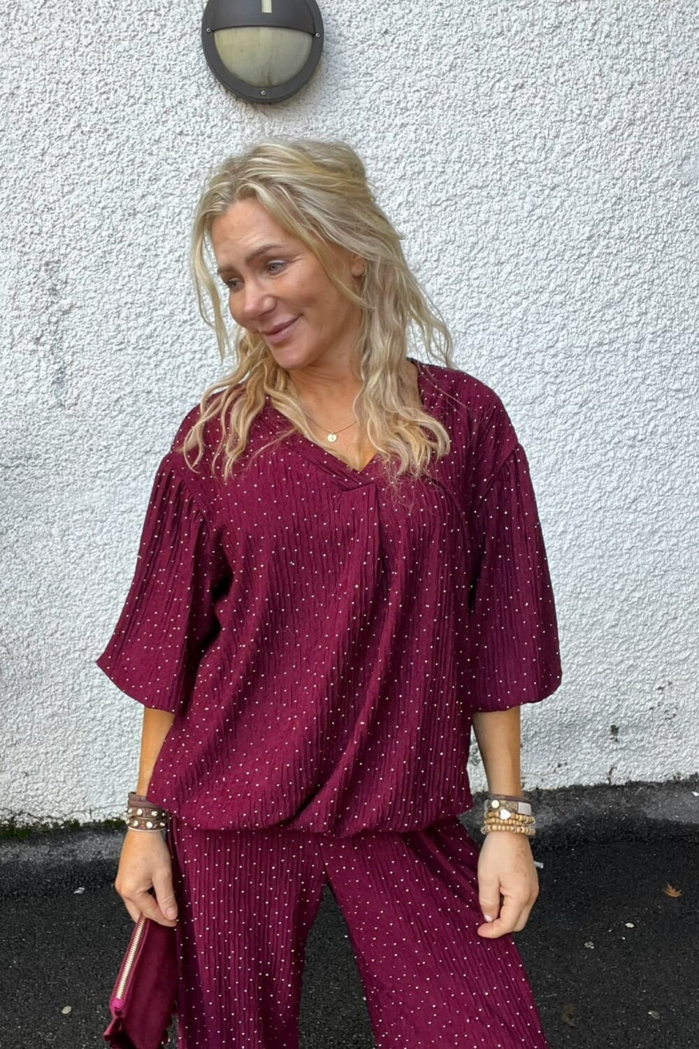 Blond kvinna står lutad mot en vit, grov putsad vägg och tittar åt sidan med ett svagt leende. Hon bär vinröd prickig blus och byxor, armband och håller en matchande clutch; rund vägglampa ovanför.