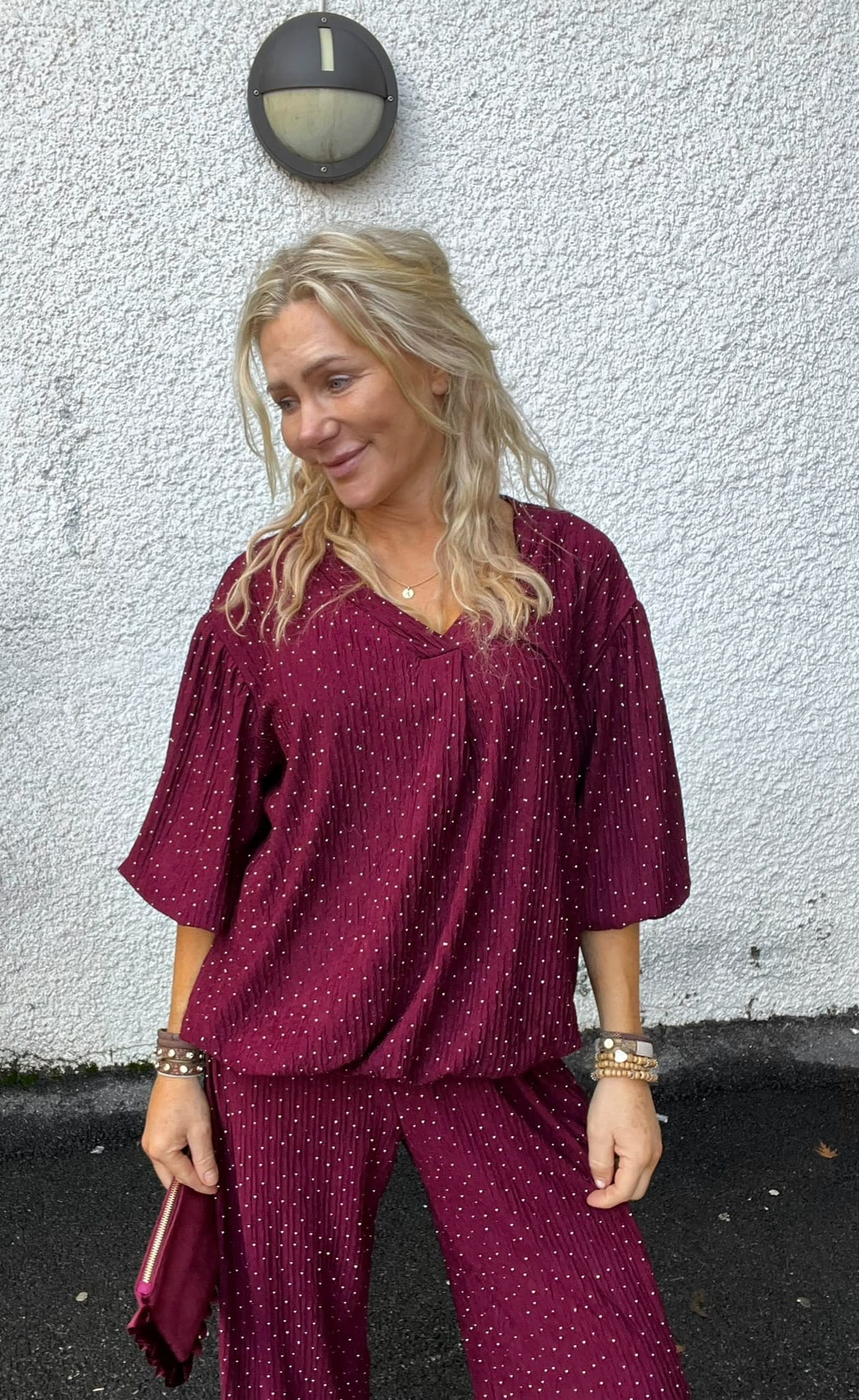 Blond kvinna står lutad mot en vit, grov putsad vägg och tittar åt sidan med ett svagt leende. Hon bär vinröd prickig blus och byxor, armband och håller en matchande clutch; rund vägglampa ovanför.