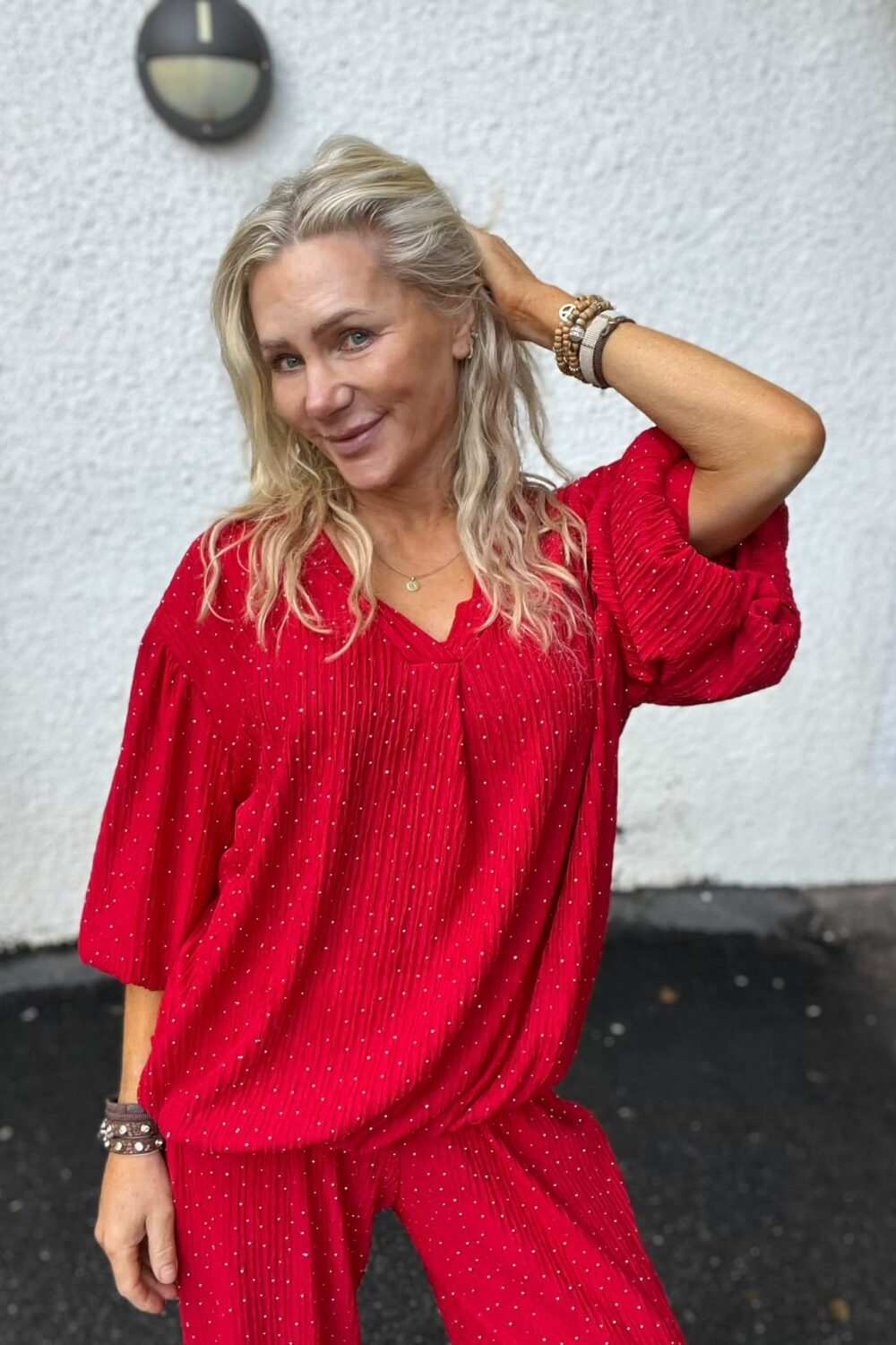 Kvinna med långt blont hår poserar och ler, med ena handen bakom huvudet. Hon bär en röd prickig jumpsuit och flera armband, och står utomhus framför en vit vägg med en rund vägglampa.