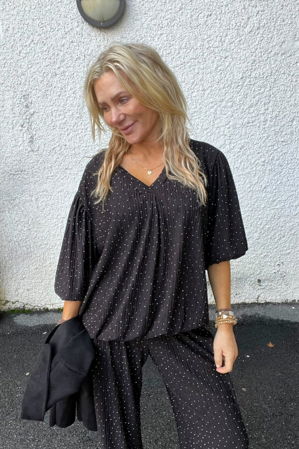 Blond kvinna i svart, prickig blus och matchande byxor står utomhus mot en vitputsad vägg och ler medan hon tittar åt sidan. Hon håller en svart jacka över armen och bär armband och halsband.