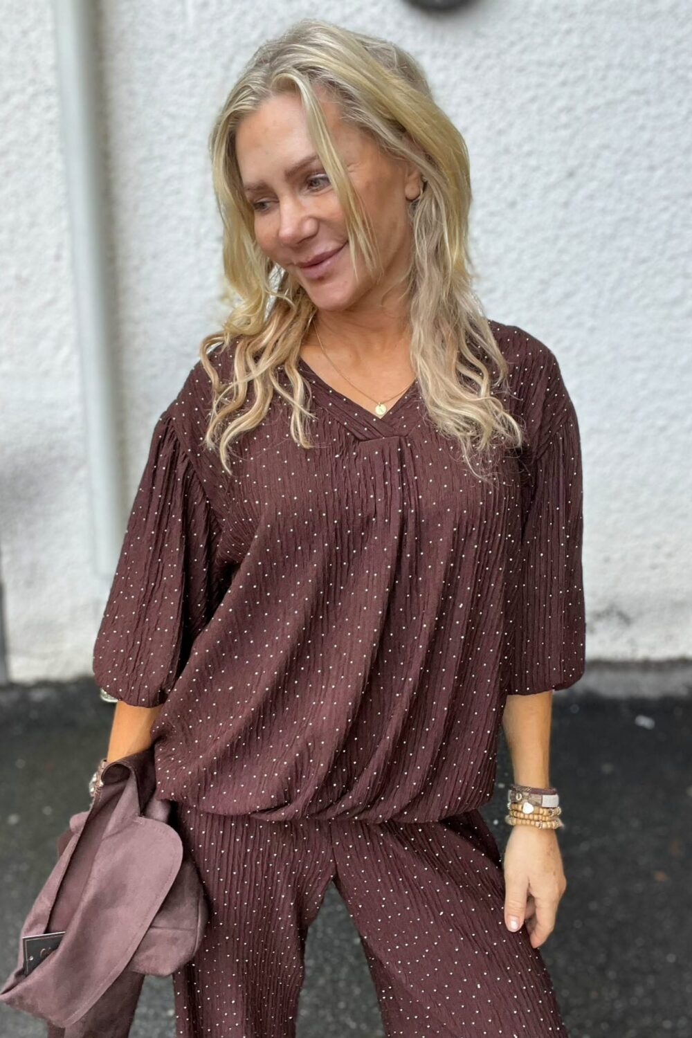 Leende blond kvinna tittar åt sidan och står avslappnat utomhus mot en ljus vägg. Hon bär brun, prickig matchande topp och byxor, halsband och armband, och håller en mauve jacka i handen.