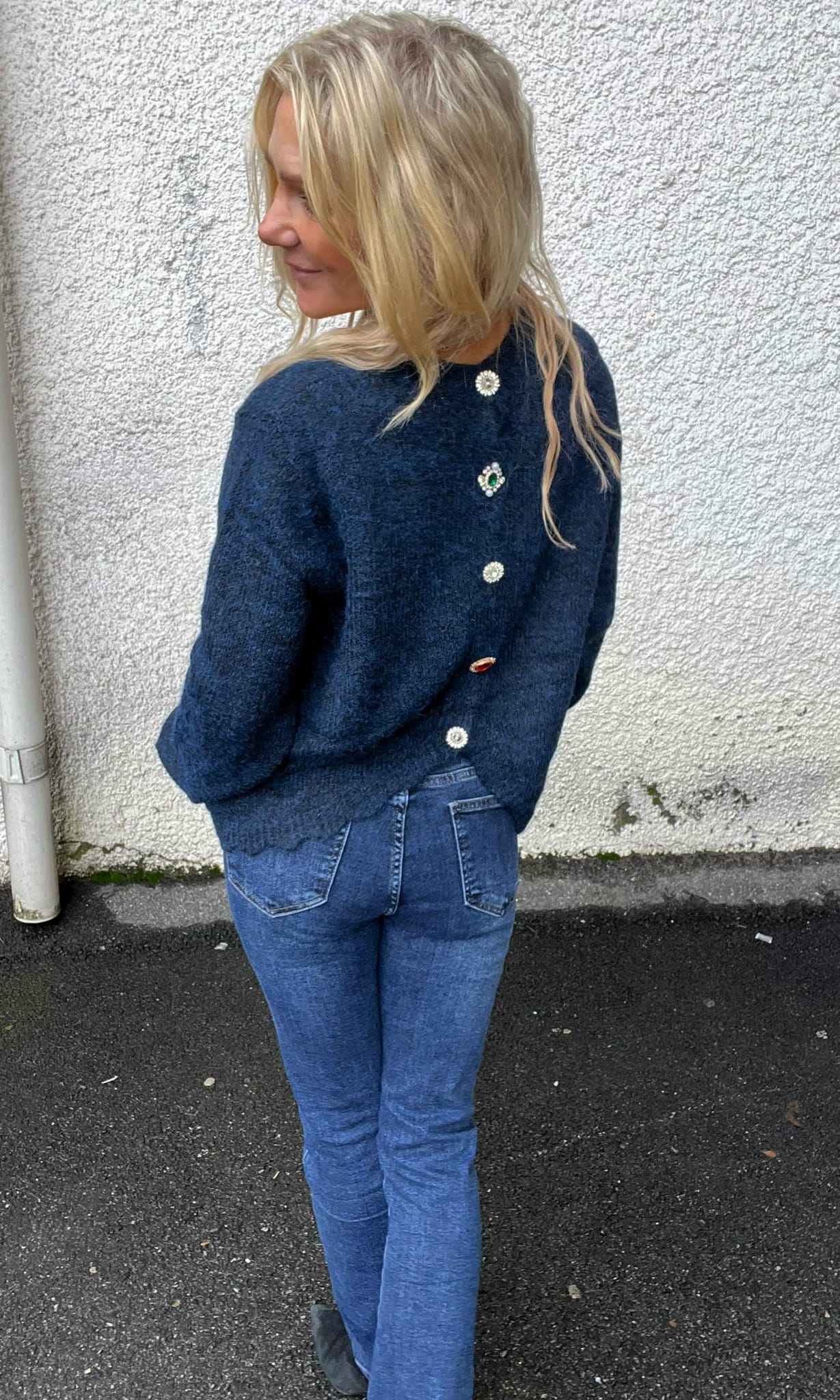 Blond kvinna står med ryggen mot kameran och tittar åt vänster, med händerna i fickorna. Hon bär blå stickad tröja med dekorativa smyckesknappar längs ryggen och jeans, vid en vit putsad vägg utomhus.