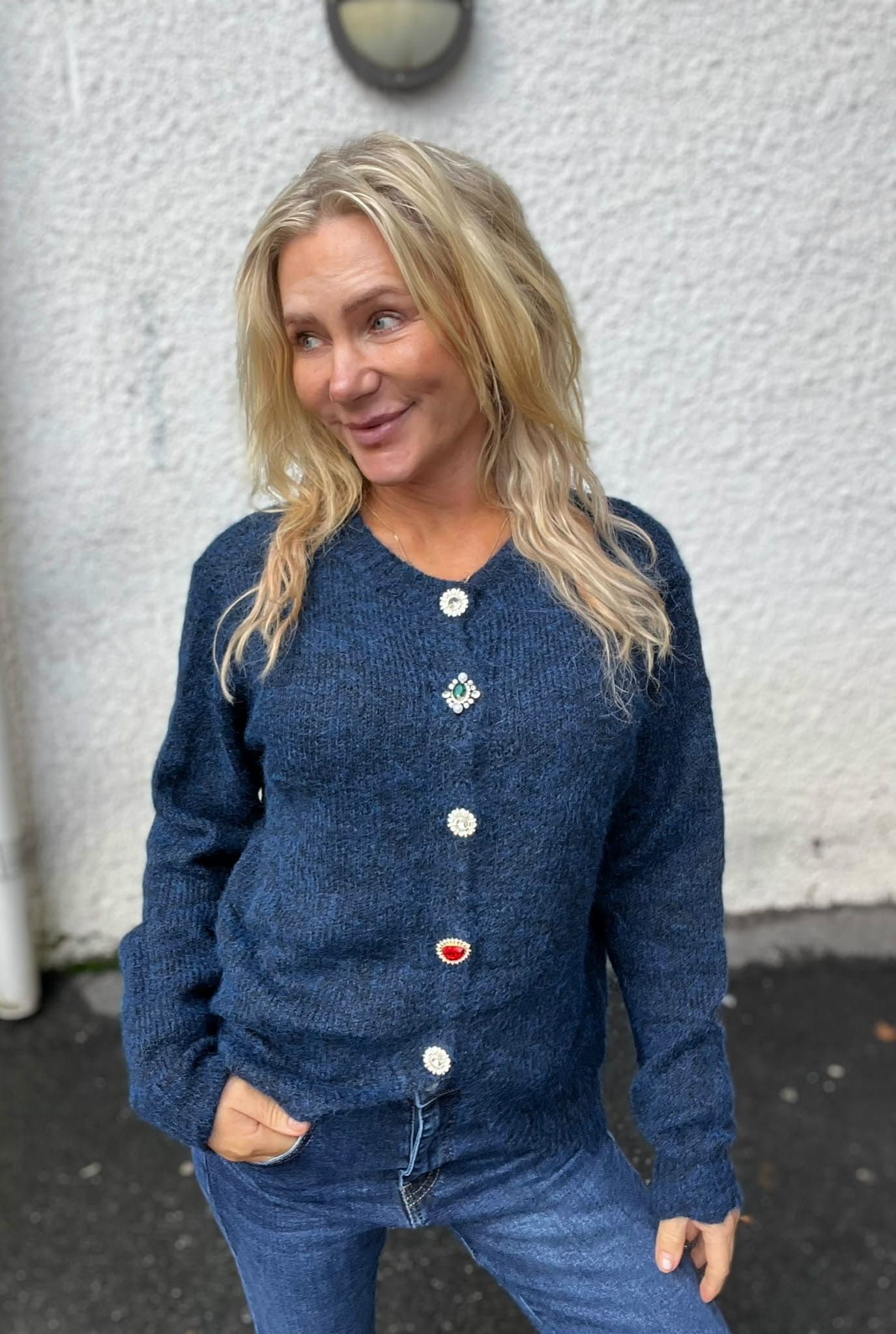 Blond kvinna står utomhus mot en ljus, putsad vägg och ler medan hon tittar åt sidan. Hon har händerna i jeansfickorna och bär en blå stickad tröja med dekorativa knappar och märken.