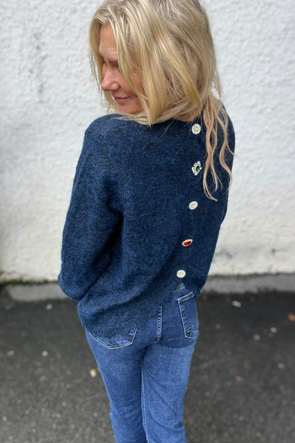 Blond kvinna står utomhus och vänder sig över axeln mot kameran, med händerna i fickorna. Hon bär mörkblå stickad tröja med dekorativa knappar och smyckesdetaljer på ryggen samt jeans, mot en ljus vägg.