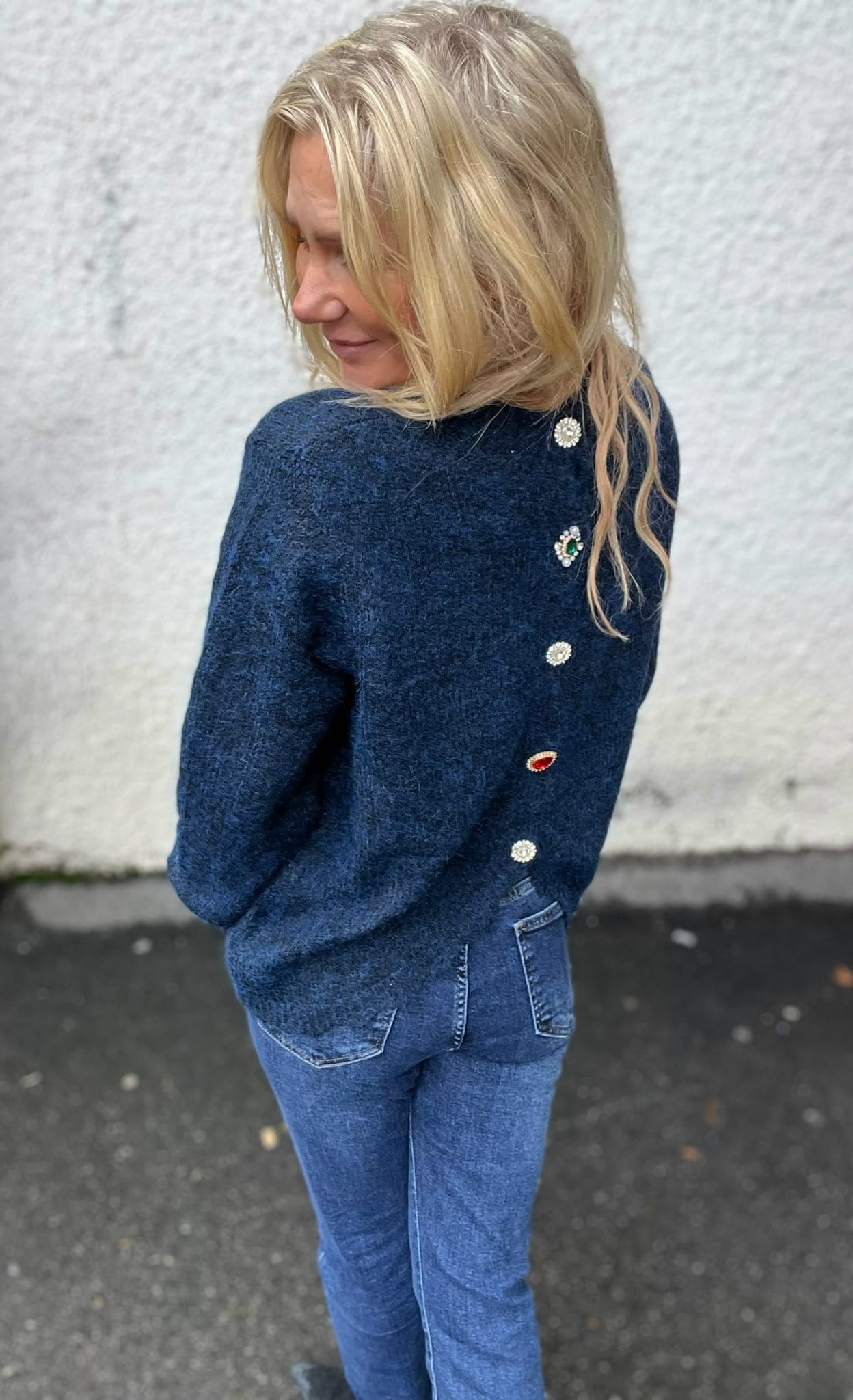 Blond kvinna står utomhus och vänder sig över axeln mot kameran, med händerna i fickorna. Hon bär mörkblå stickad tröja med dekorativa knappar och smyckesdetaljer på ryggen samt jeans, mot en ljus vägg.