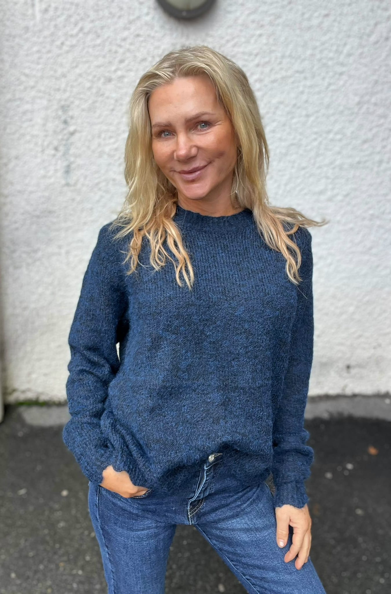 Blond kvinna i mörkblå stickad tröja och jeans står utomhus med händerna i fickorna och ler mot kameran. Bakgrunden är en ljus, grovputsad vägg och våt asfalt.