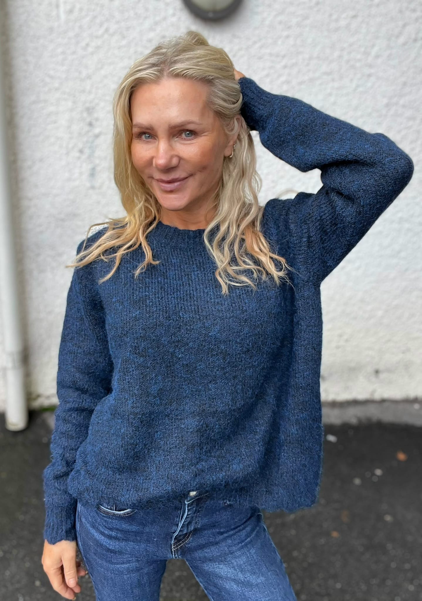 Blond kvinna i blå stickad tröja och jeans poserar utomhus, ler mot kameran och håller ena handen bakom huvudet. Bakom henne syns en ljus, grov vägg och mörk asfalt.