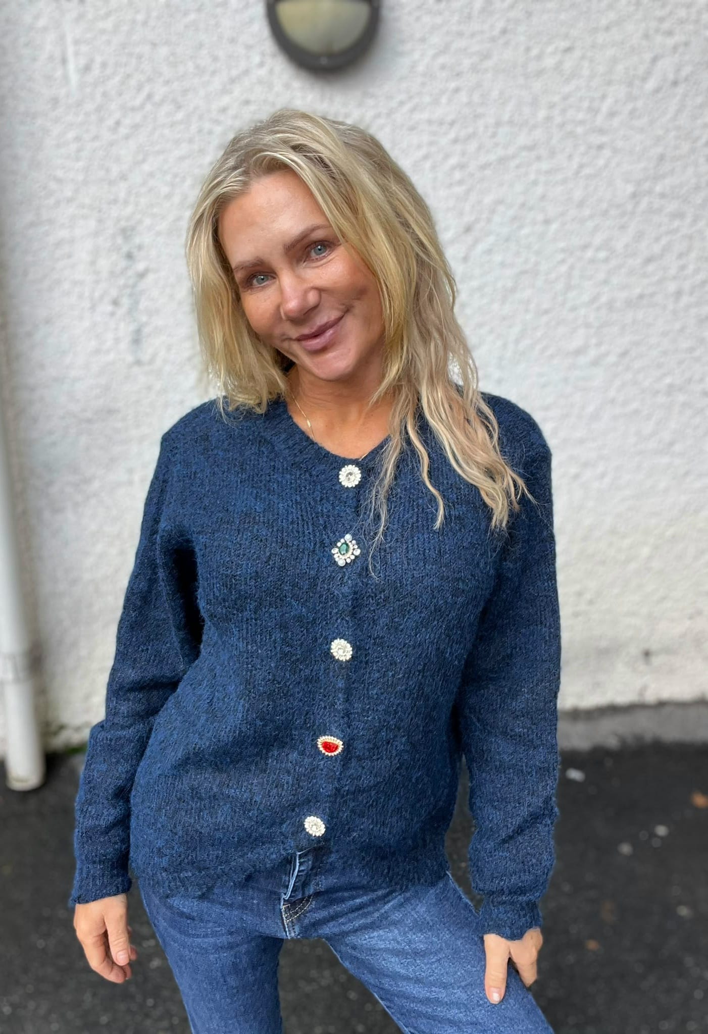 Leende kvinna med långt blont hår står utomhus och lutar sig lätt åt sidan framför en ljus putsad vägg. Hon bär blå stickad kofta med dekorativa smyckesknappar och blå jeans.