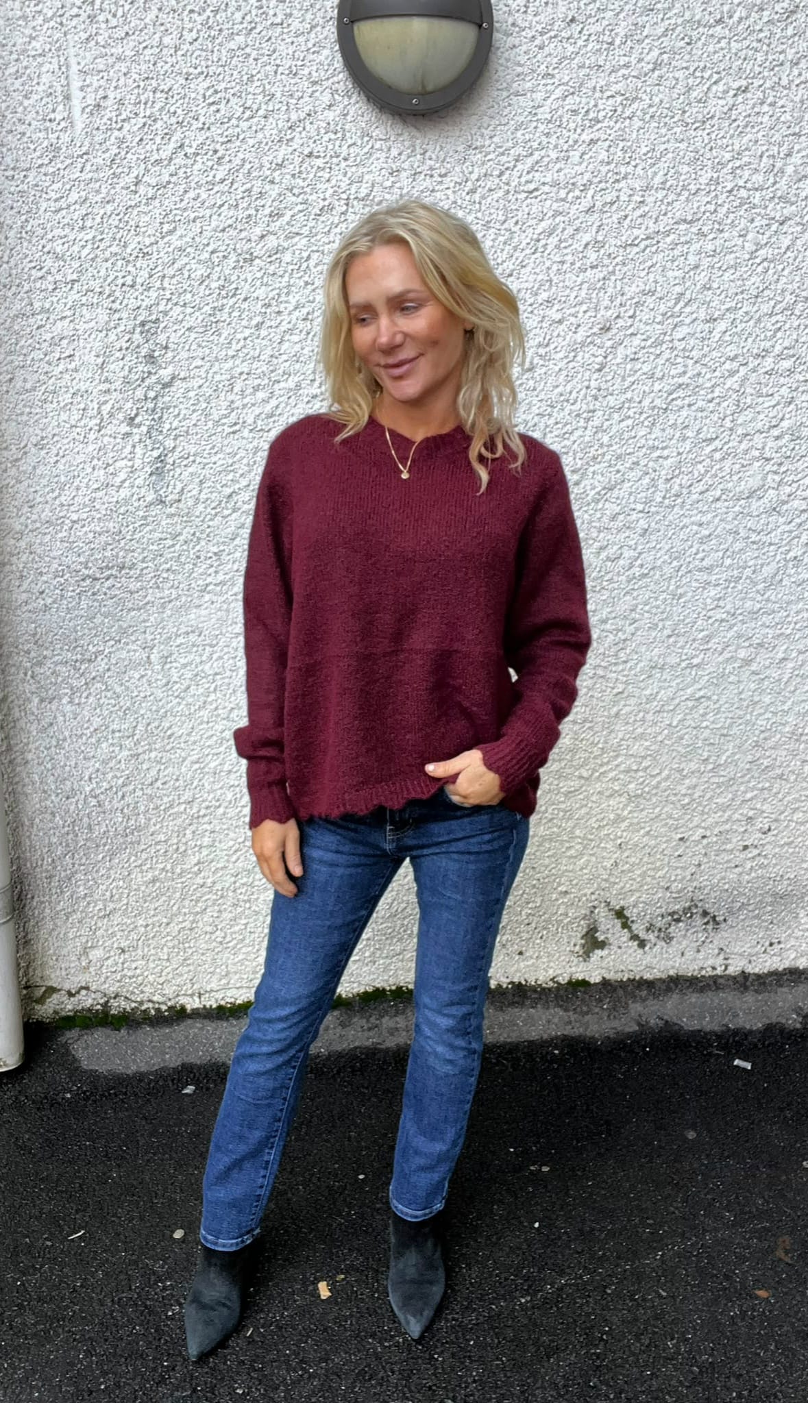 Blond kvinna står avslappnat med ena handen i jeansfickan och tittar åt sidan, klädd i vinröd stickad tröja, blå jeans och svarta boots. Hon poserar utomhus mot en vit putsad vägg under en rund vägglampa.