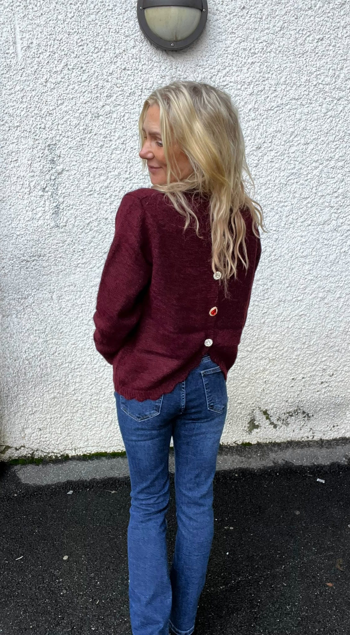 Blond kvinna står med ryggen delvis mot kameran och tittar över axeln, klädd i vinröd stickad tröja med dekorativa knappar bak och blå jeans, framför en vit putsad vägg med rund vägglampa.