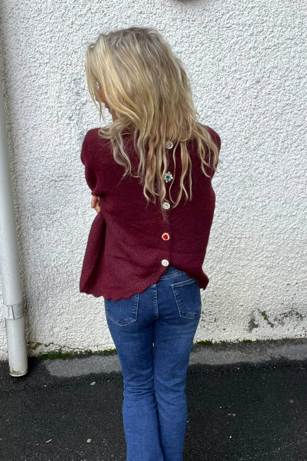 Person med långt blont, vågigt hår står med ryggen mot kameran och armarna korsade, lutad mot en vit putsad vägg. Bär vinröd tröja med dekorativa knappar på ryggen och blå jeans; vägglampa ovanför.