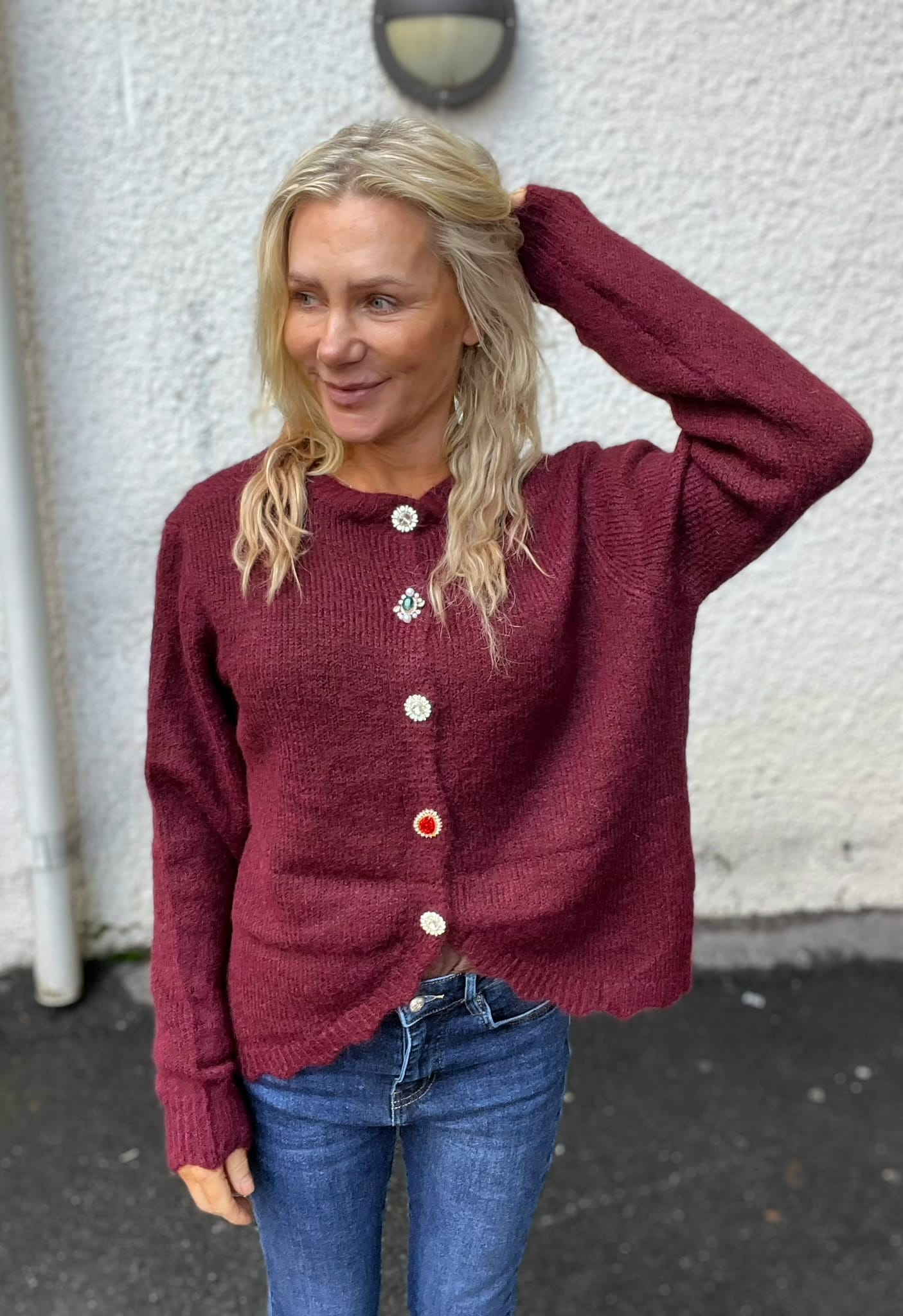 Blond kvinna står utomhus mot en ljus, grov vägg och ler medan hon håller ena handen bakom huvudet. Hon bär vinröd stickad kofta med dekorativa knappar och blå jeans; en vägglampa syns ovanför.