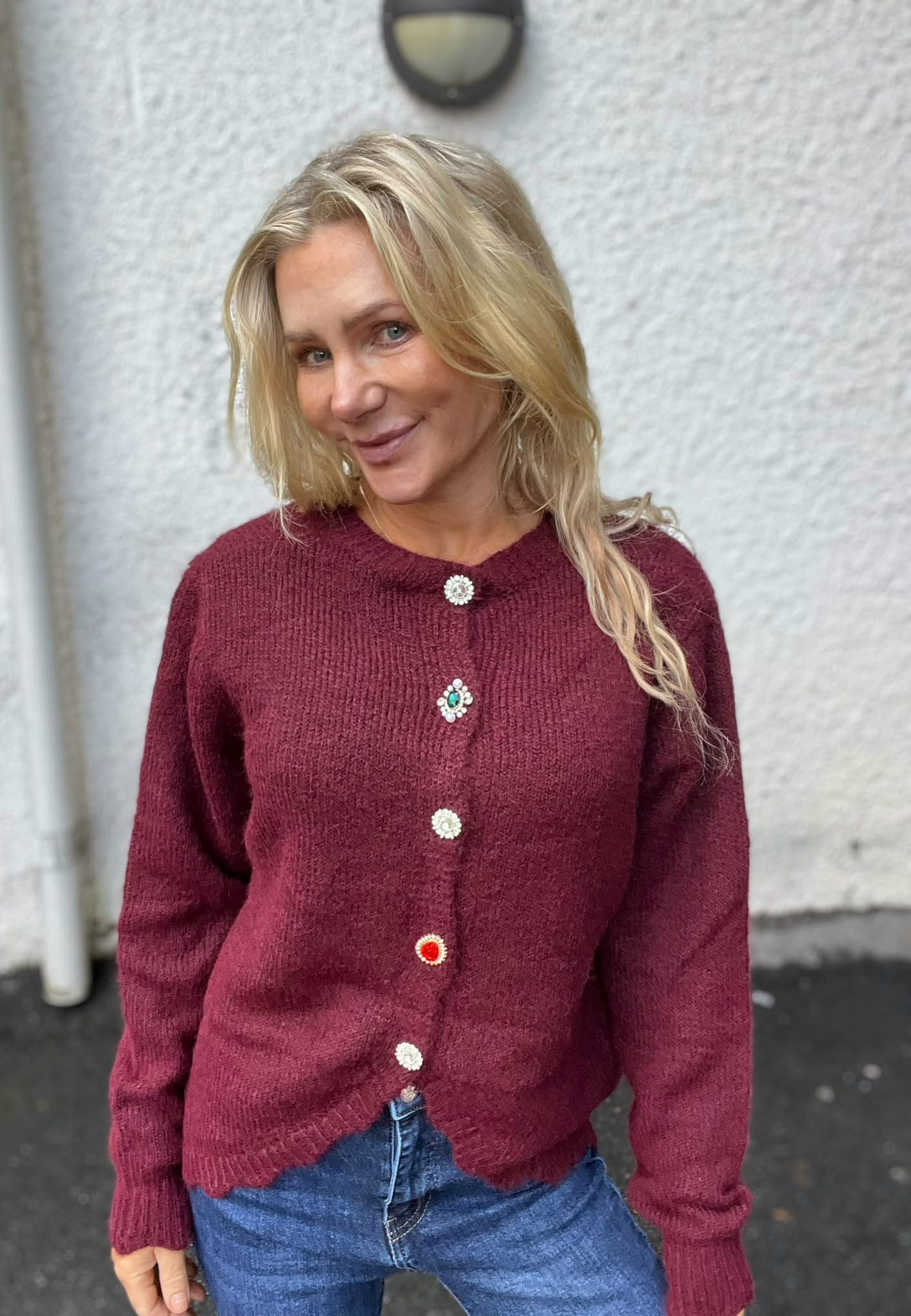 Blond kvinna poserar och ler lätt mot kameran framför en ljus, grov vägg utomhus. Hon bär en vinröd stickad kofta med dekorativa, färgade knappar och blå jeans, med håret över ena axeln.