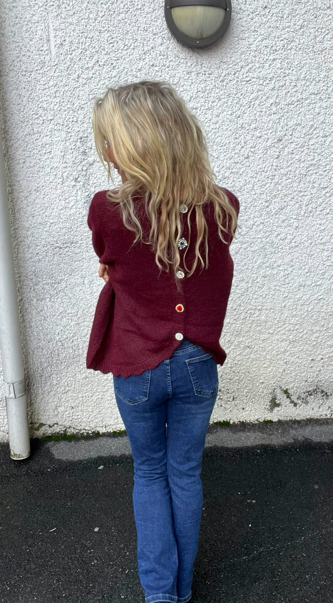 Person med långt blont, vågigt hår står med ryggen mot kameran och armarna korsade, lutad mot en vit putsad vägg. Bär vinröd tröja med dekorativa knappar på ryggen och blå jeans; vägglampa ovanför.