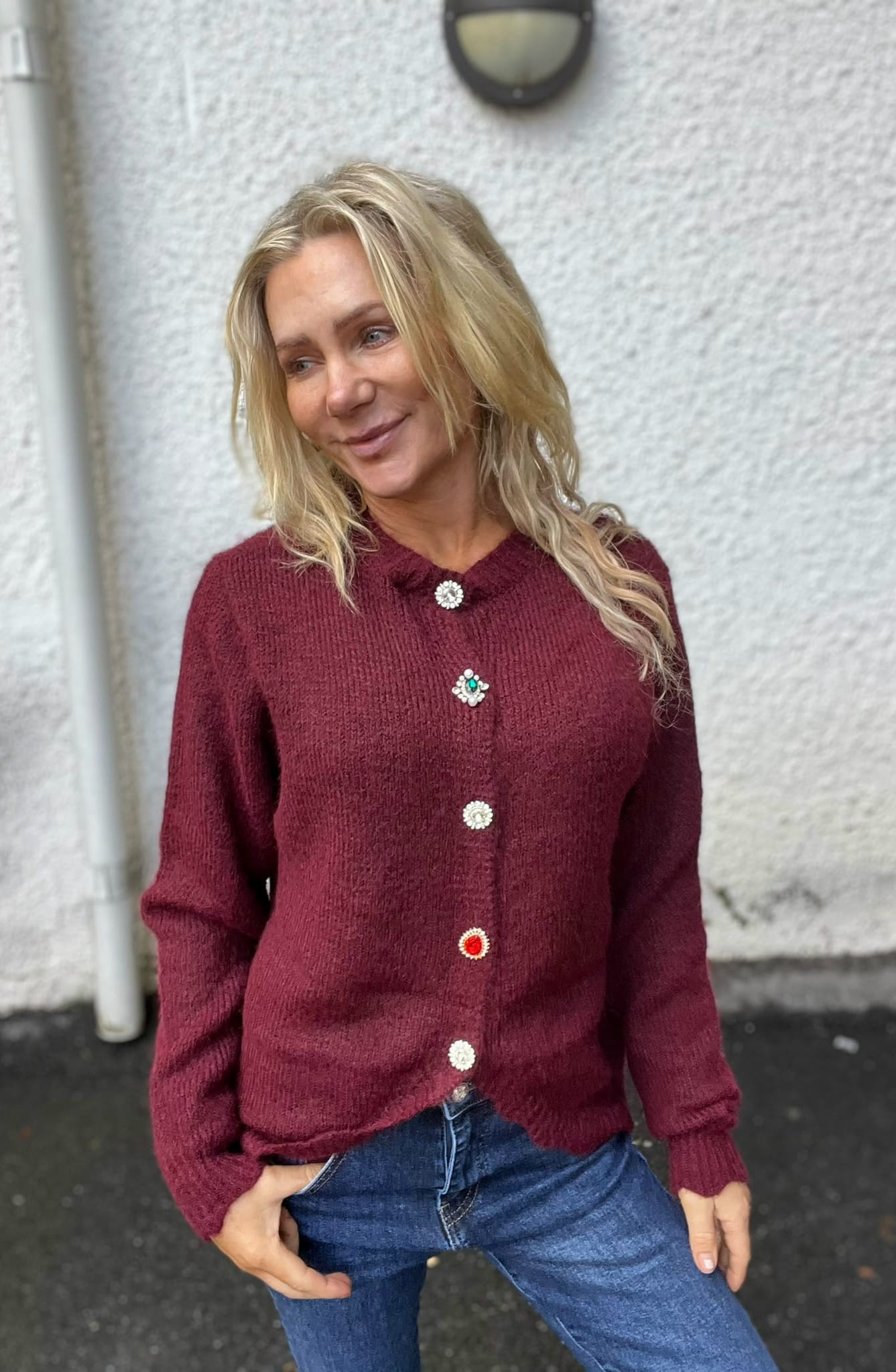 Blond kvinna står lutad mot en ljus putsad vägg och ler medan hon tittar åt sidan. Hon bär en vinröd stickad kofta med dekorativa, färgade knappar och blå jeans, händerna i fickorna.