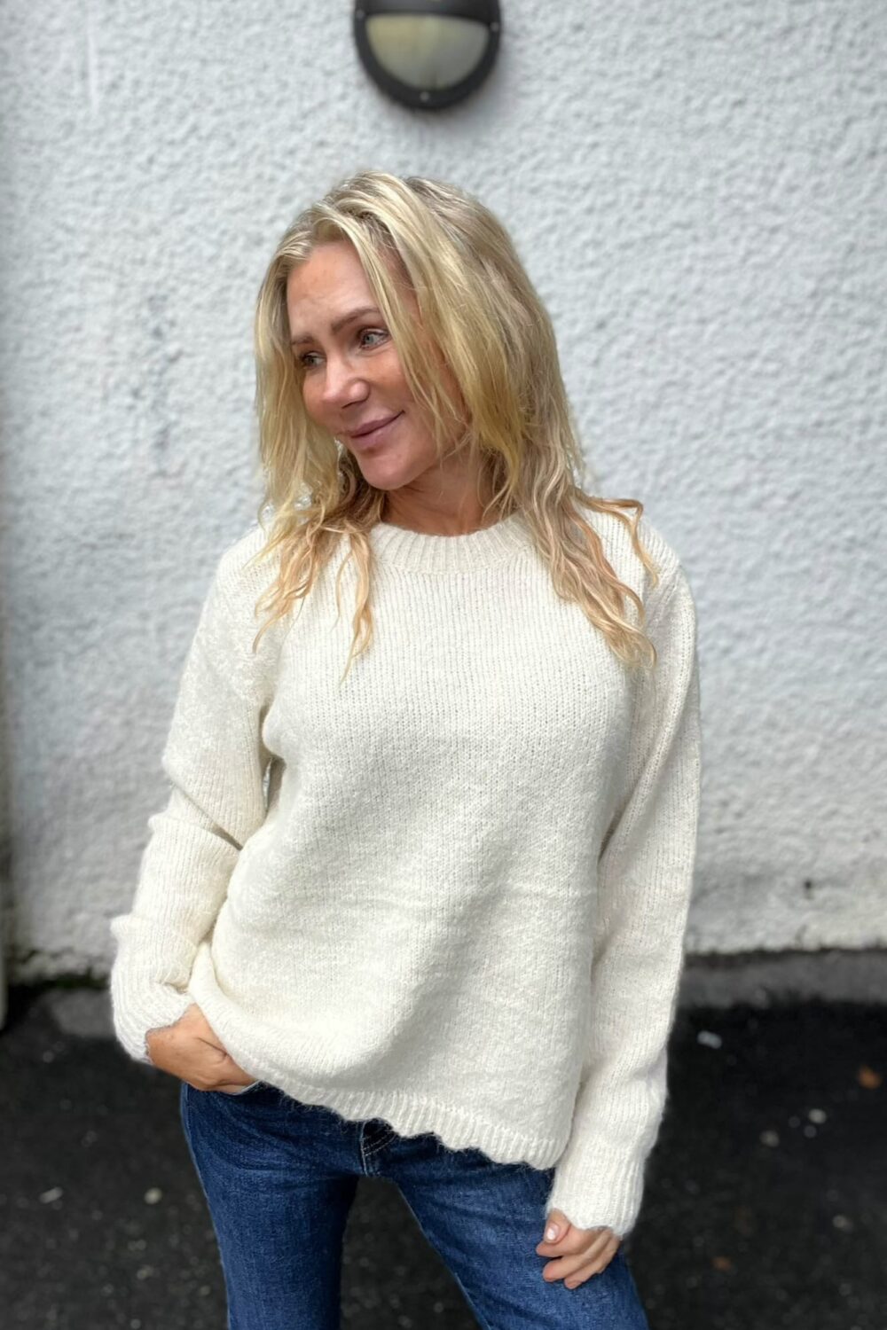 Blond kvinna i vit stickad tröja och blå jeans står avslappnat med en hand i fickan och ler svagt åt sidan, framför en ljusgrå putsad vägg med rund vägglampa ovanför.