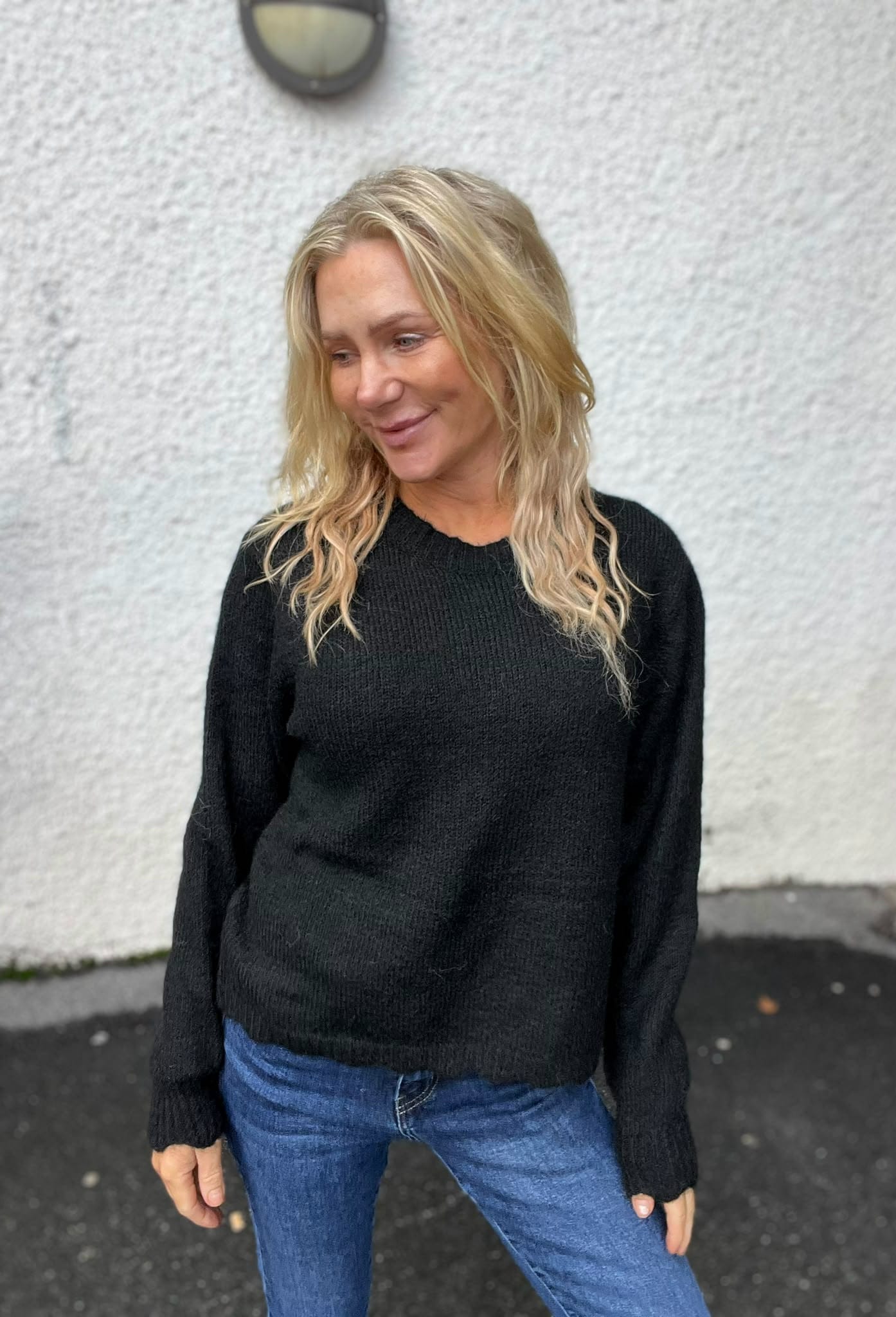 Blond kvinna i svart stickad tröja och blå jeans står utomhus framför en ljus putsad vägg och ler medan hon tittar åt sidan. Markytan är mörk asfalt, och en rund vägglampa syns ovanför.