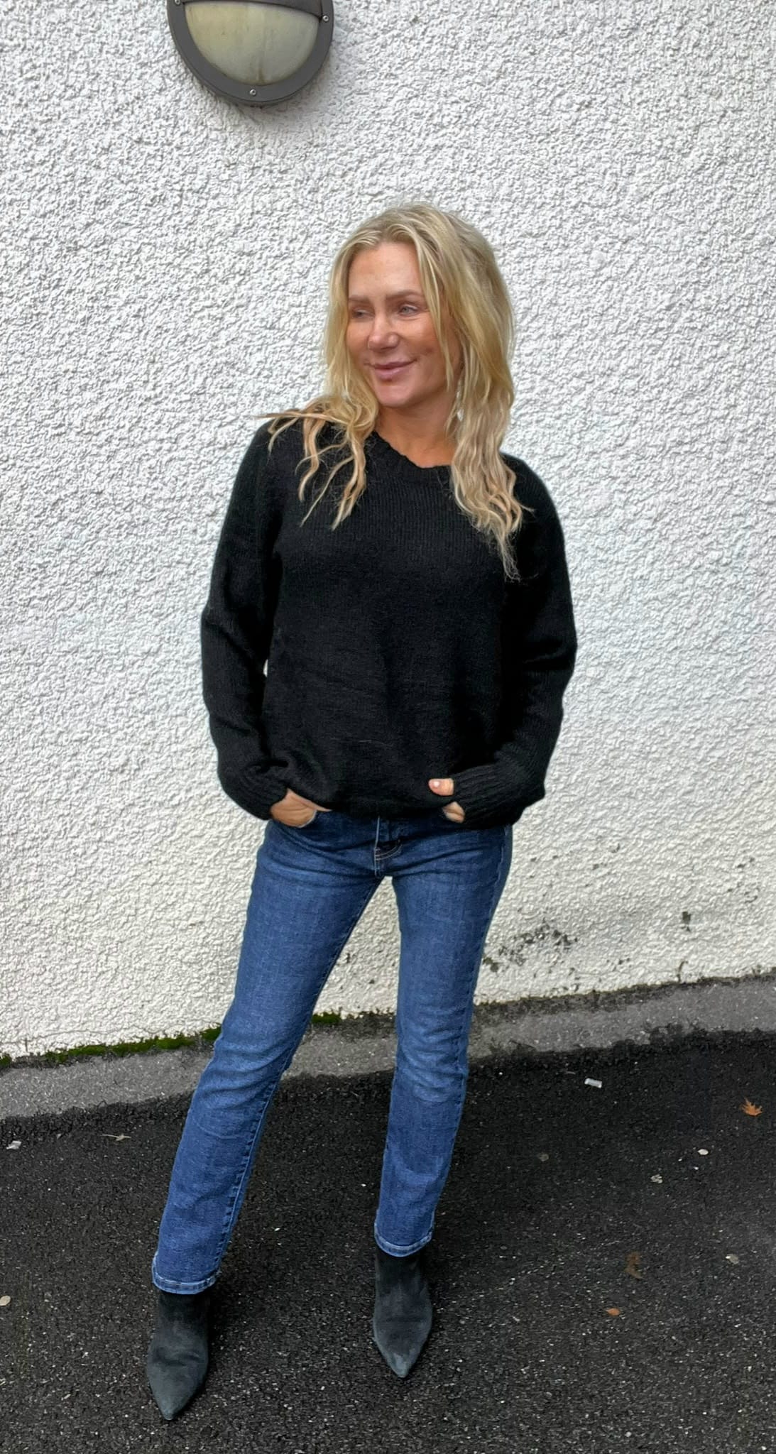 Blond kvinna står avslappnat med händerna i fickorna och ler svagt, iklädd svart stickad tröja, blå jeans och svarta boots. Hon poserar utomhus mot en vit, grovputsad vägg med rund vägglampa ovanför.