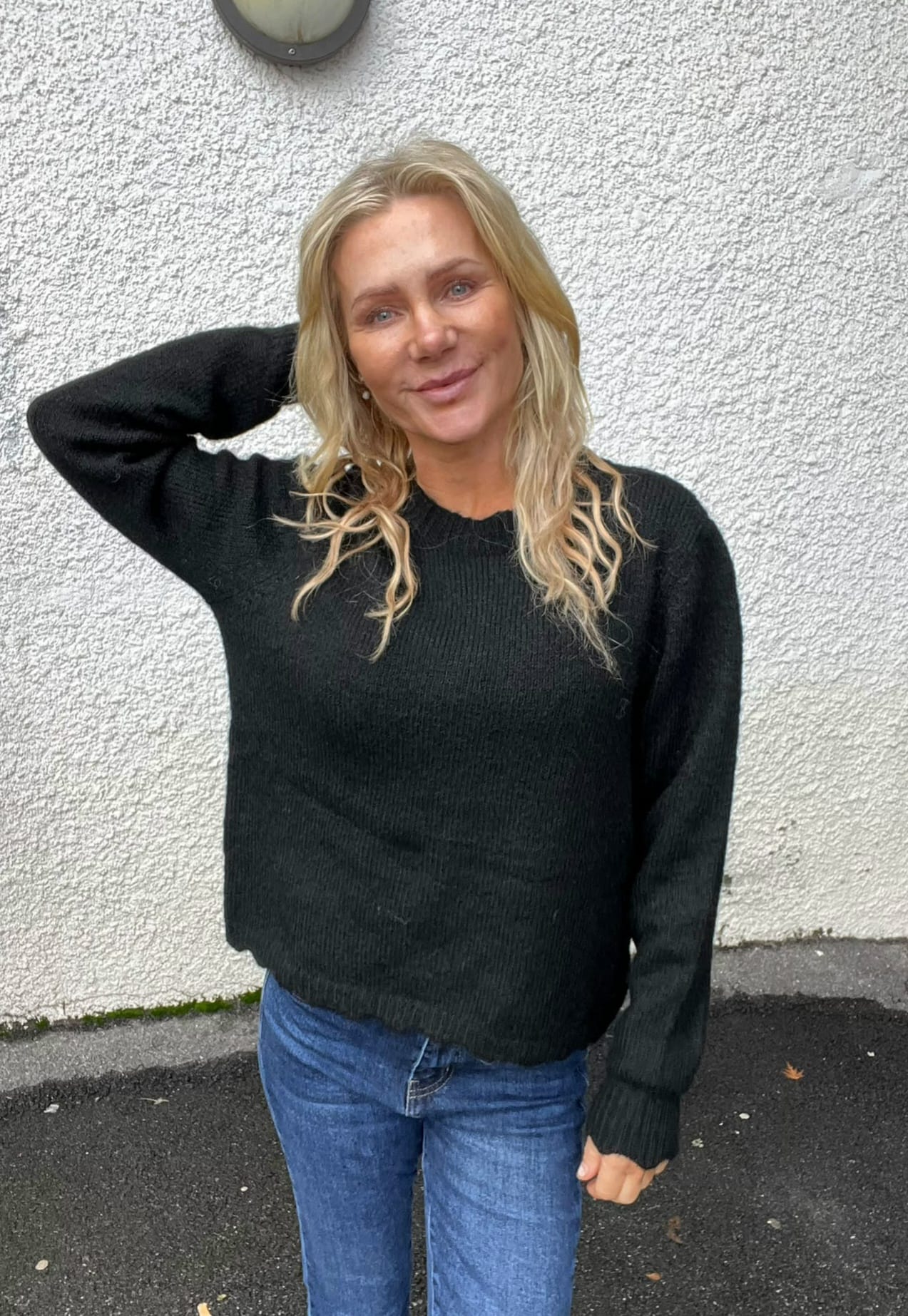 Blond kvinna står och ler mot kameran med ena handen bakom huvudet, klädd i mörk stickad tröja och blå jeans. Hon står utomhus framför en ljus, grovputsad vägg med en rund vägglampa ovanför.