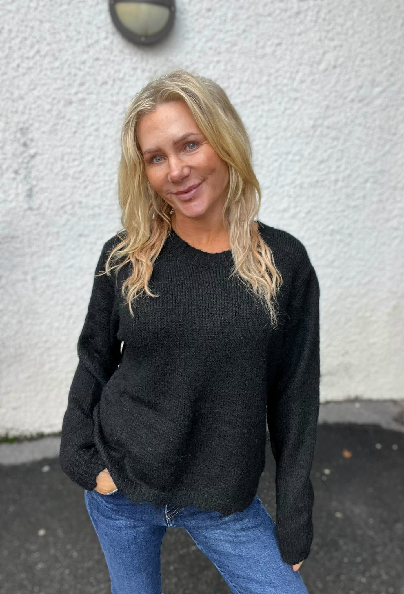 Blond kvinna ler mot kameran och står lätt lutad med händerna i jeansfickorna. Hon bär svart stickad tröja och blå jeans, fotograferad utomhus framför en ljus, grovputsad vägg med en rund vägglampa ovanför.