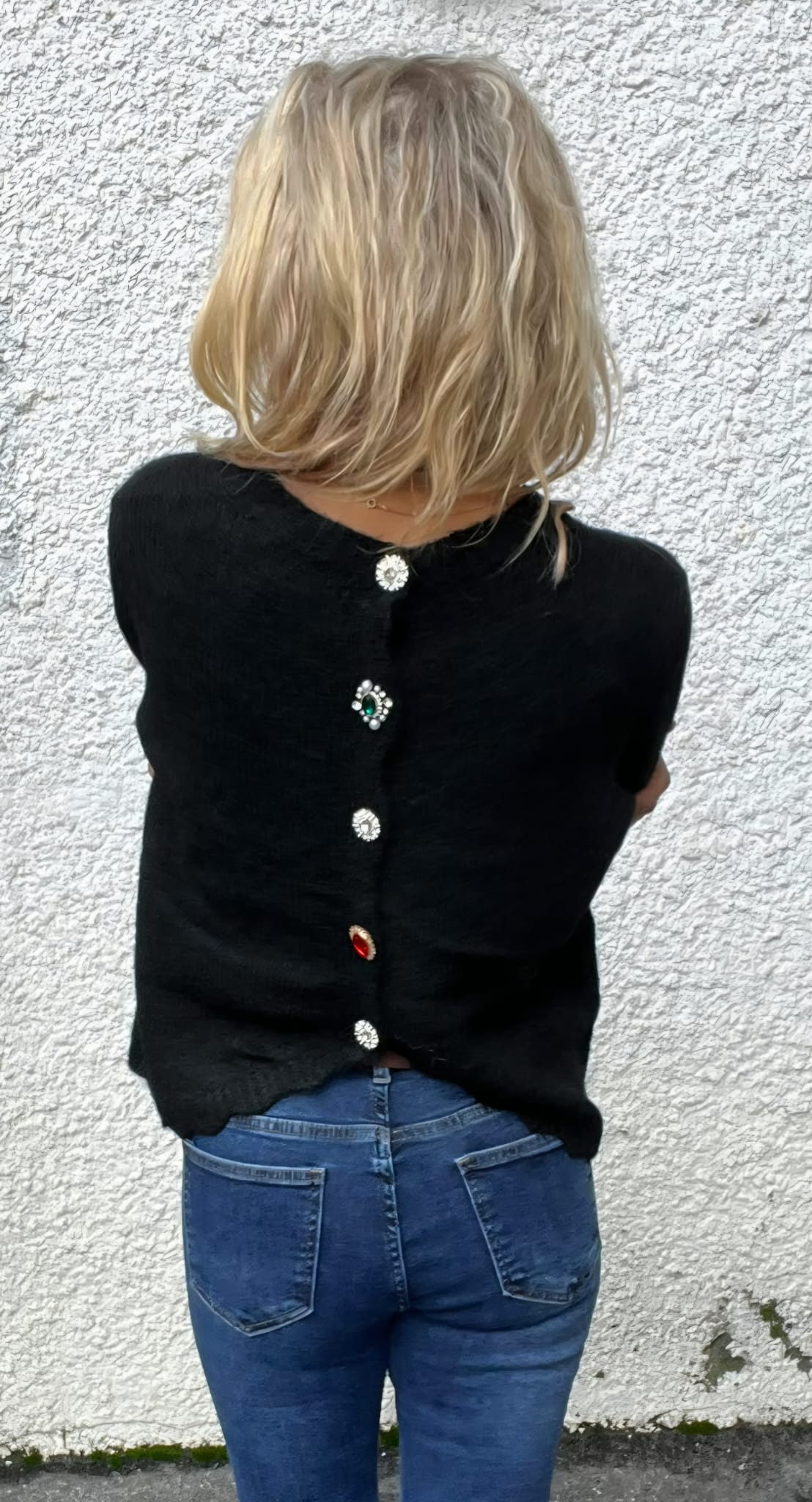 Person med axellångt blont hår står med ryggen mot kameran och armarna korsade. Hen bär blå jeans och en svart kofta med dekorativa, glittrande knappar längs ryggen, mot en vit, grovputsad vägg.