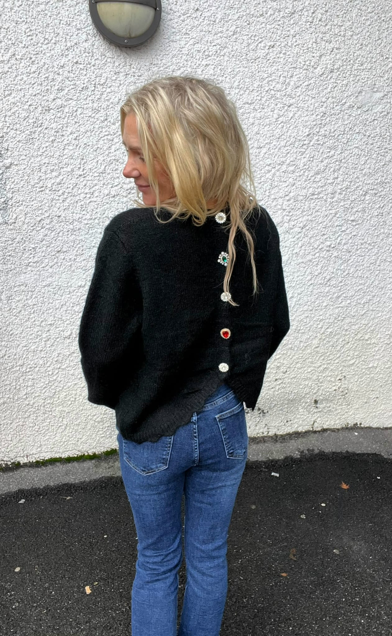 Kvinna med blont axellångt hår står i profil och tittar åt vänster. Hon bär svart stickad tröja med dekorativa knappar i ryggen och blå jeans, framför en vit putsad vägg med vägglampa.