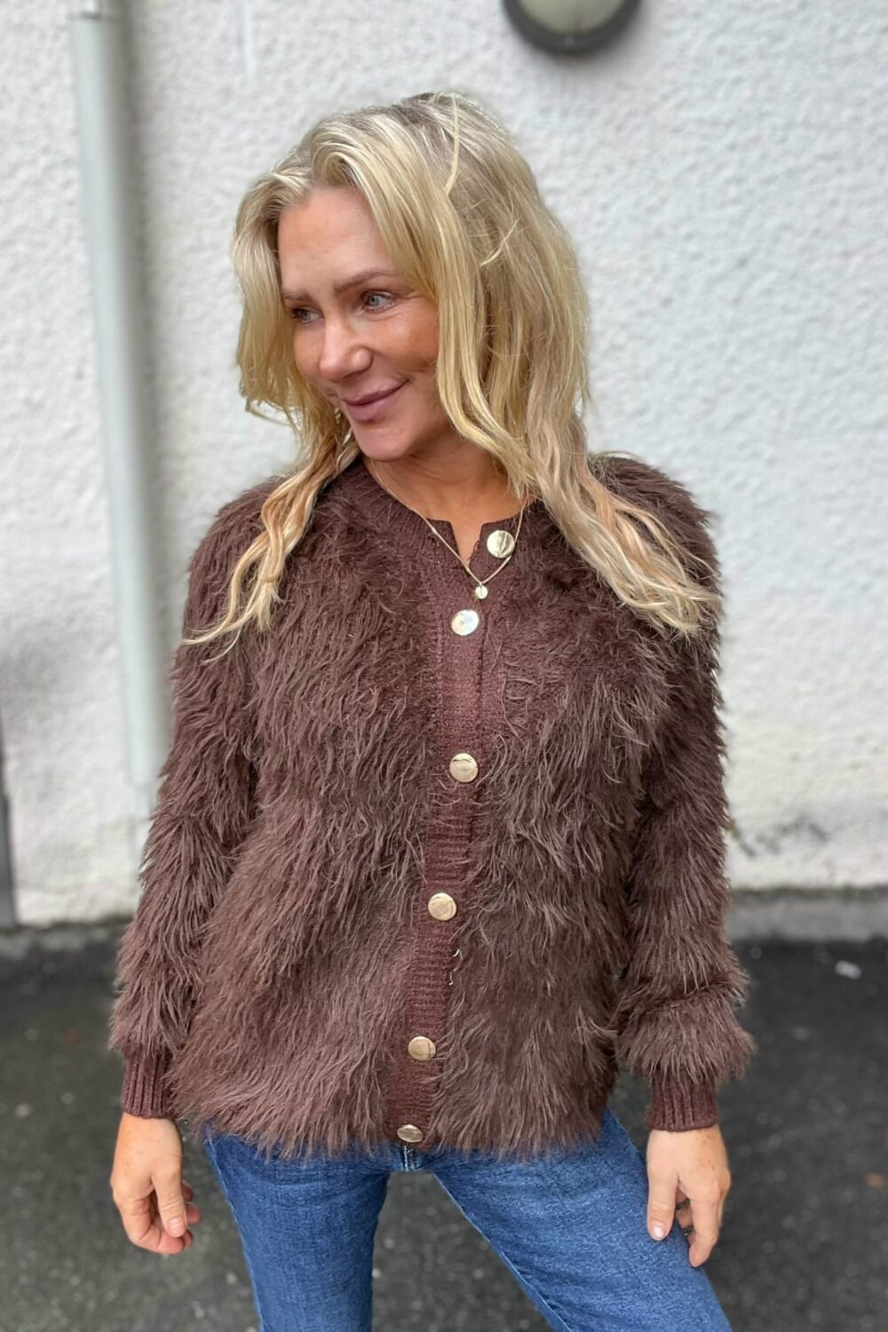 Blond kvinna står utomhus och ler medan hon tittar åt sidan. Hon bär en brun, lurvig kofta med stora knappar över en brun topp, halsband med rund berlock och blå jeans, mot en ljus vägg.